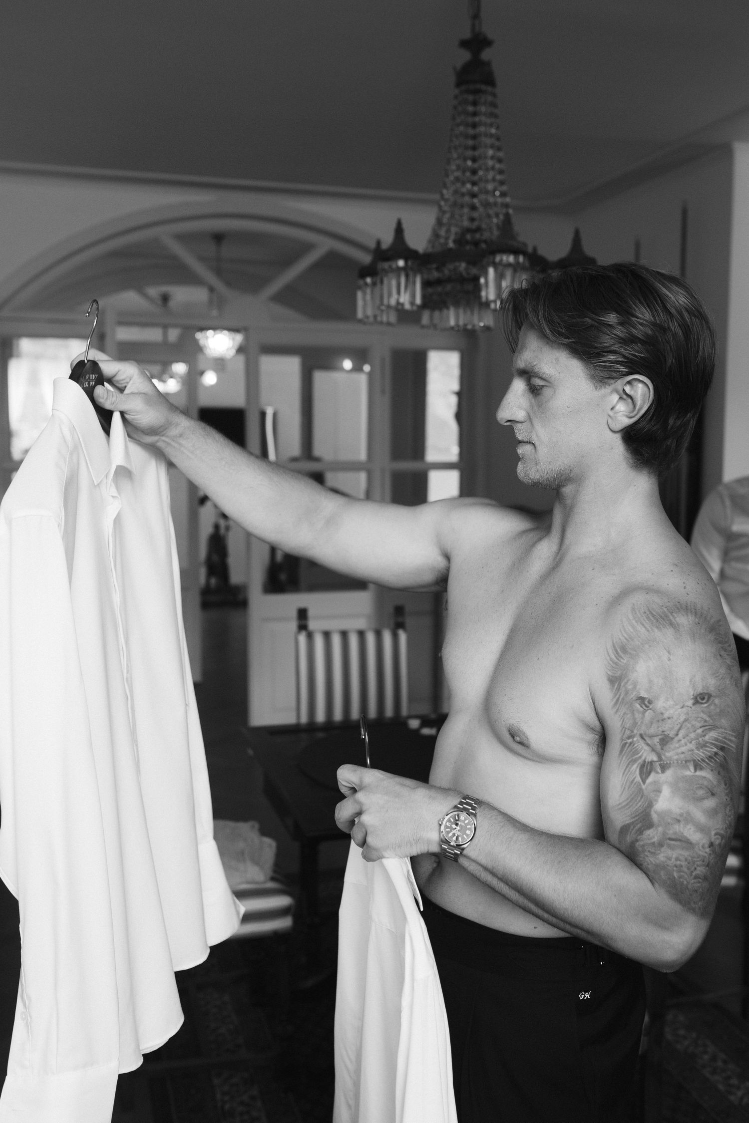 Shirtless tattooed man holding white dress shirt on hanger in elegant room with chandelier, black and white photo.