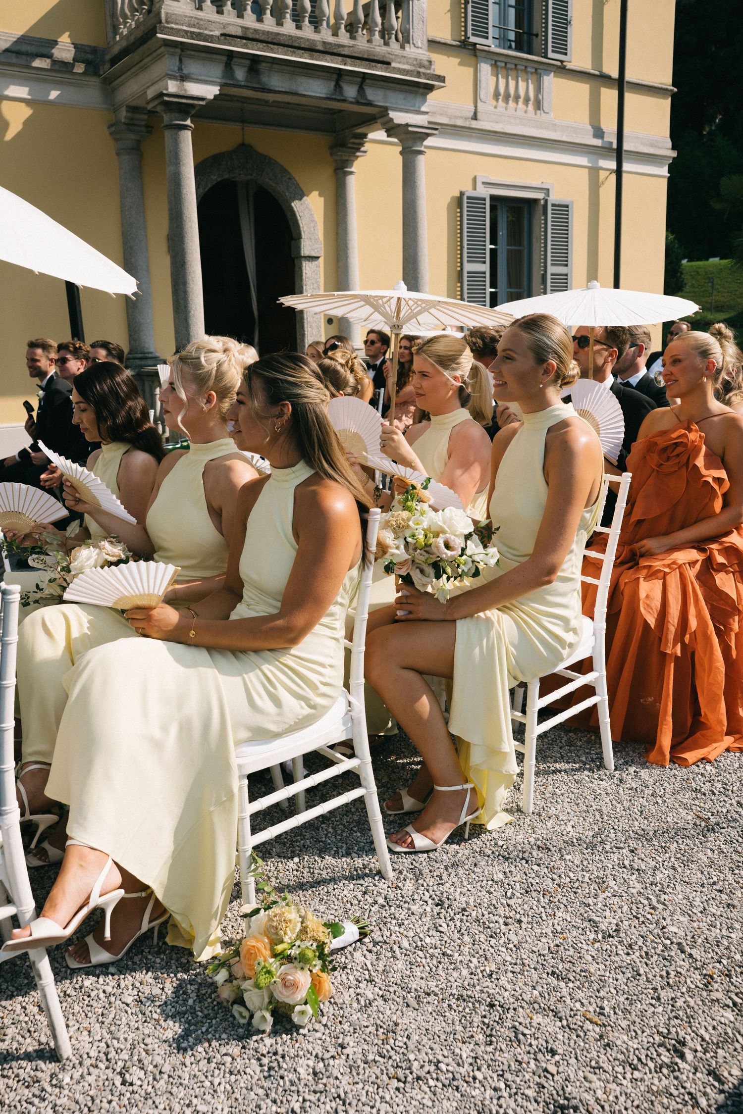 Bridesmaids in yellow dresses seated outdoors at an elegant Italian villa wedding ceremony on a sunny day.