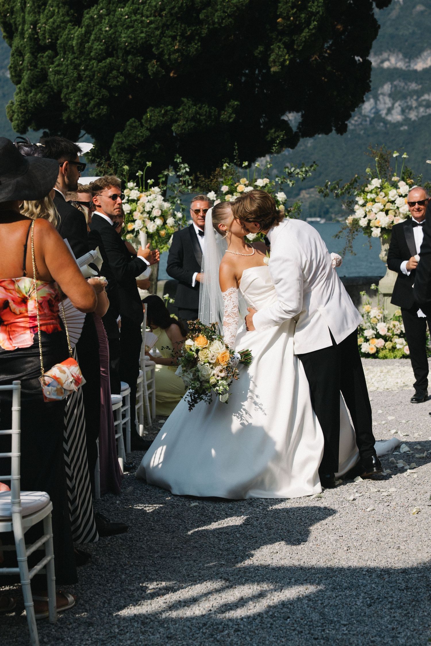 Bride and groom share a kiss at outdoor wedding ceremony with floral arch and guests watching.