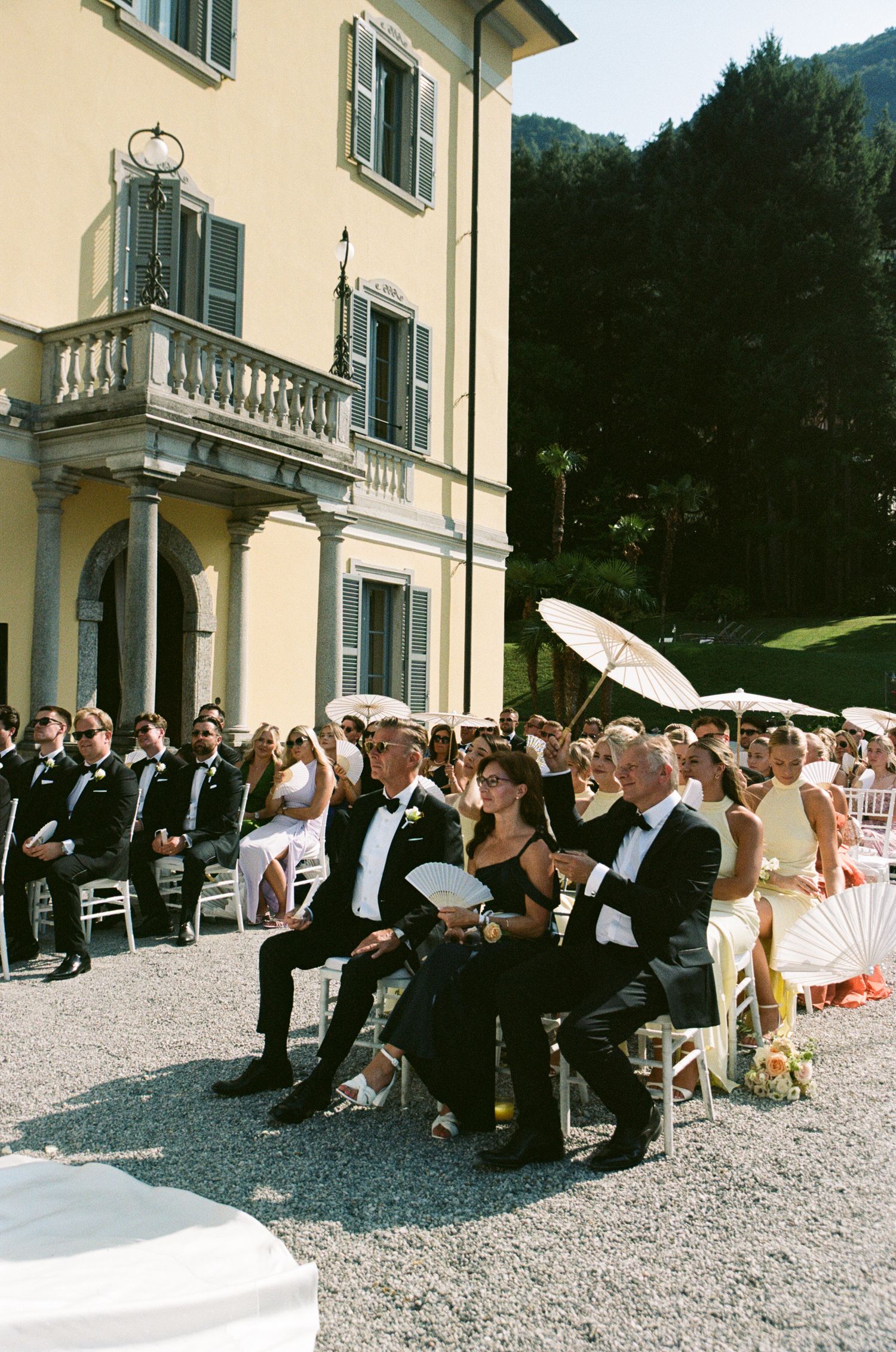 Guests in formal attire seated outdoors at an elegant Italian villa wedding ceremony on a sunny day.