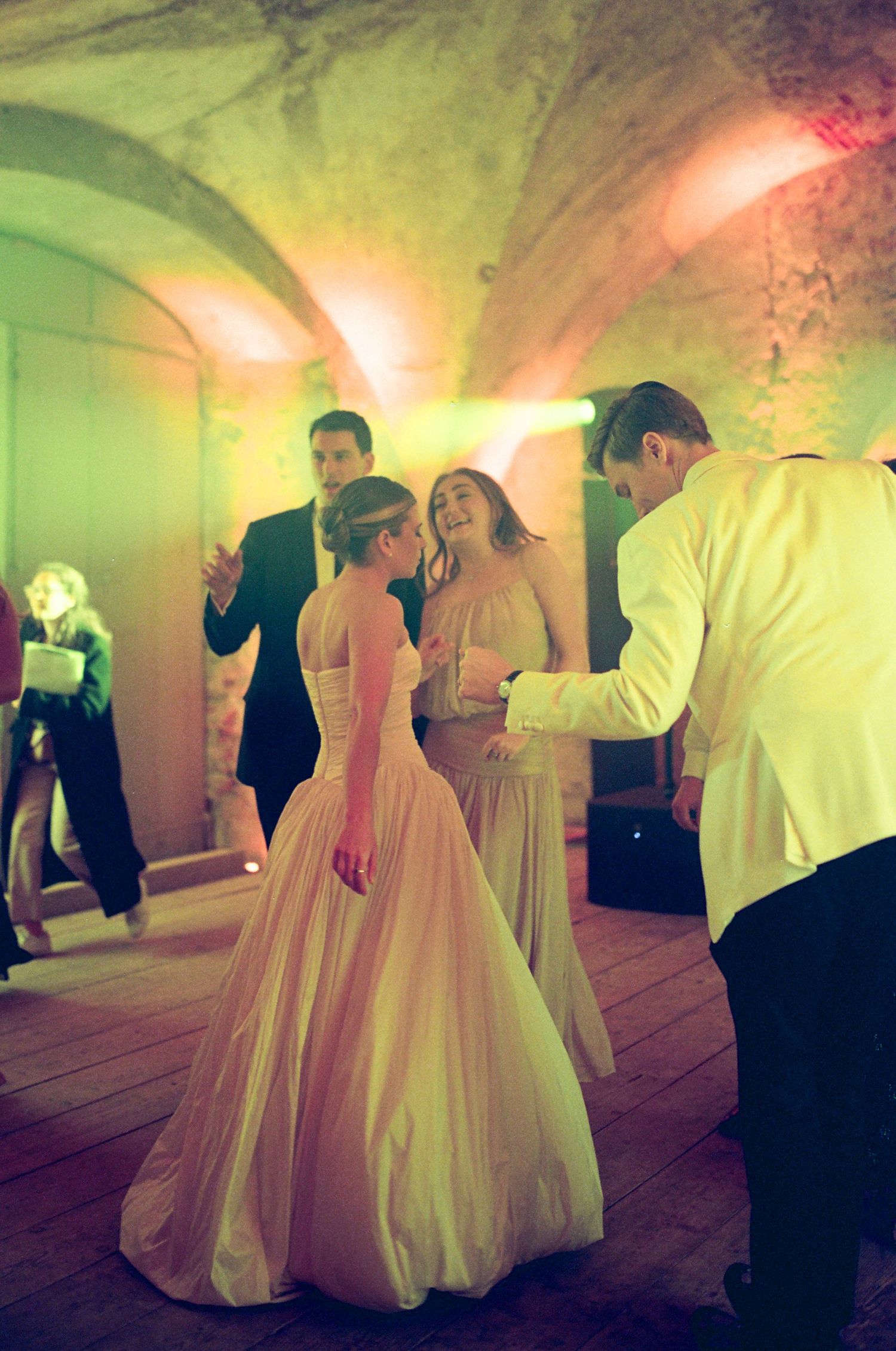 Elegant wedding guests dancing in a vaulted stone venue with warm green and pink lighting.