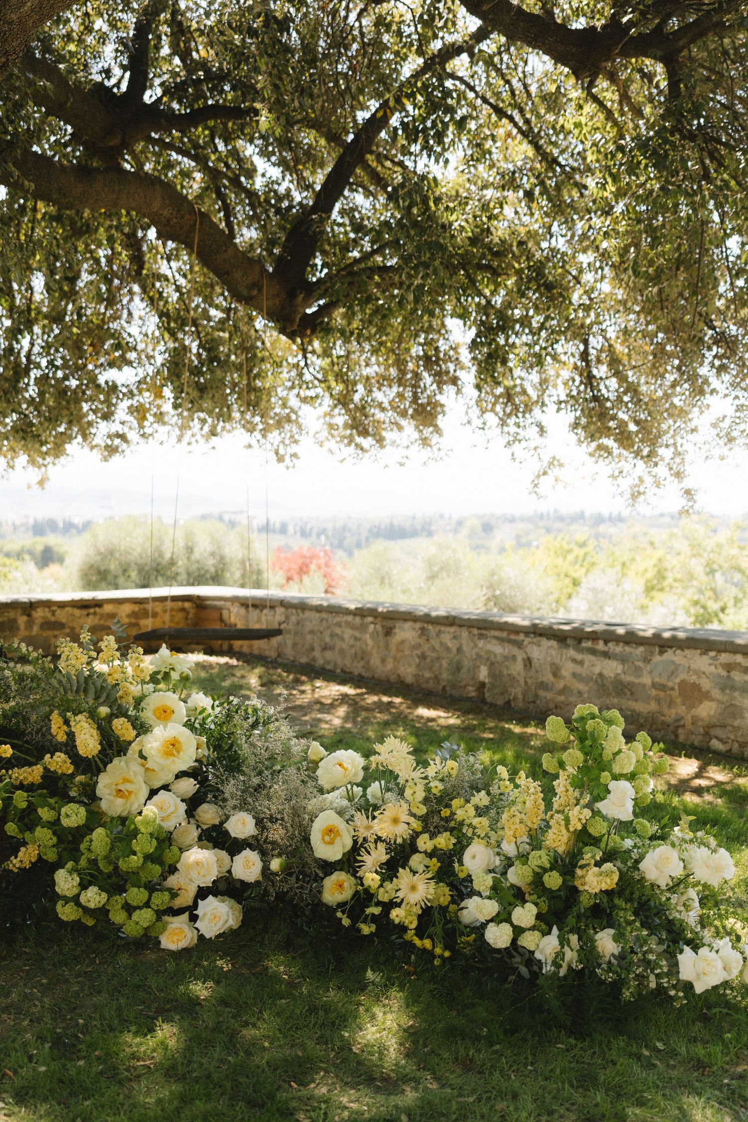 Lush white and yellow floral arrangements on grass beneath a sprawling oak tree with a scenic countryside view.