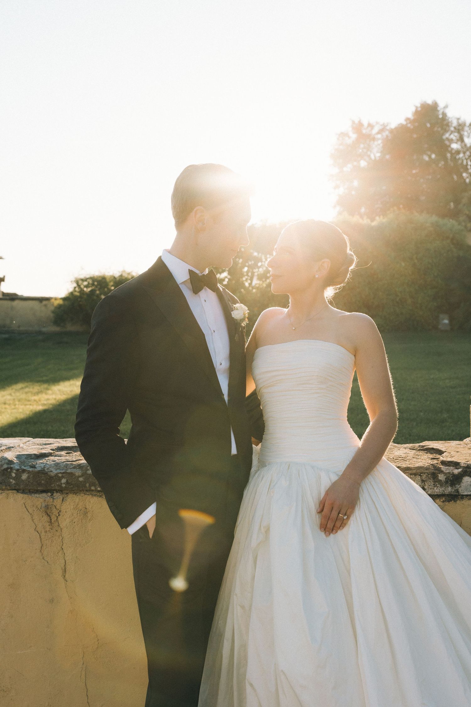 Bride and groom share a romantic moment at golden hour, sun glowing behind them near a stone wall outdoors.