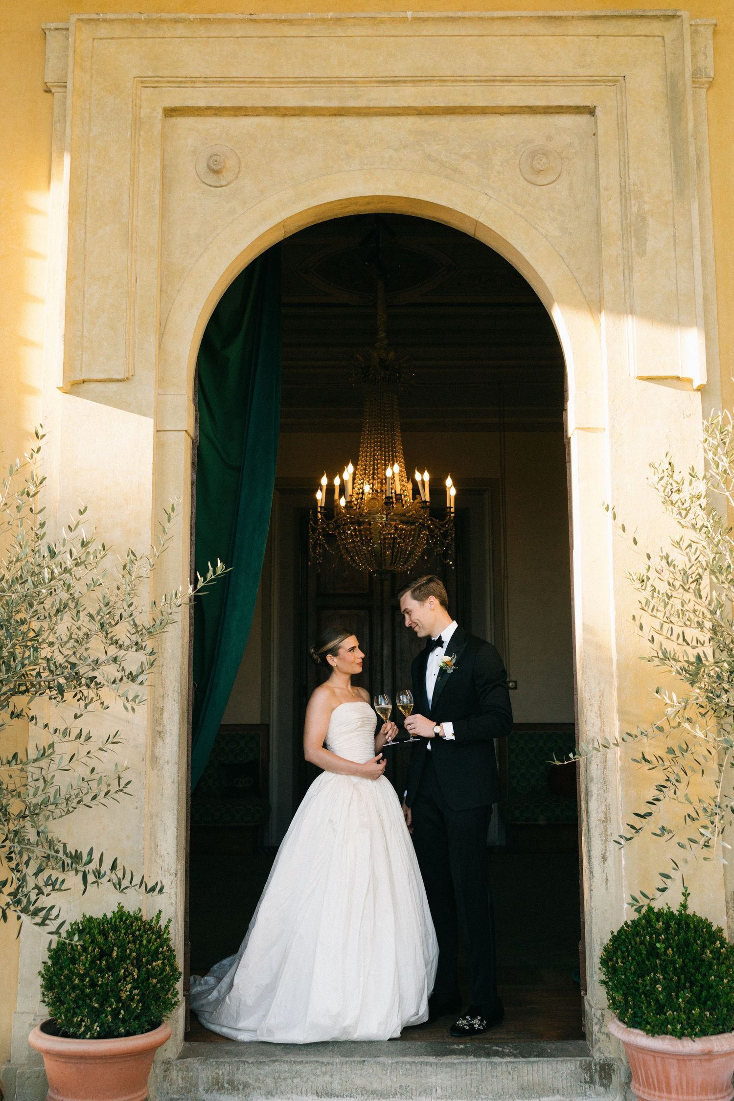 Bride and groom toasting with champagne in an elegant arched doorway with chandelier backdrop.