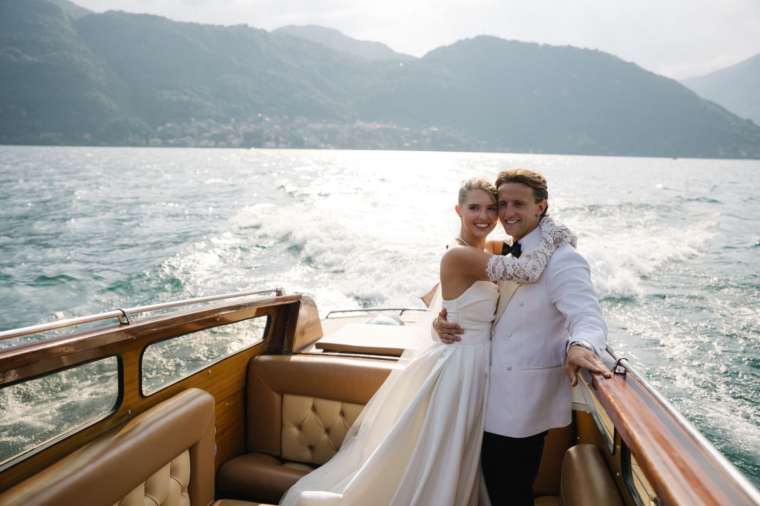 Bride and groom embrace on classic wooden speedboat on Lake Como with mountain backdrop.