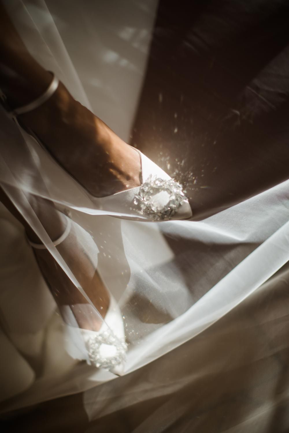 Close-up of bride's white satin wedding shoes adorned with delicate floral embellishments beneath a flowing bridal gown.