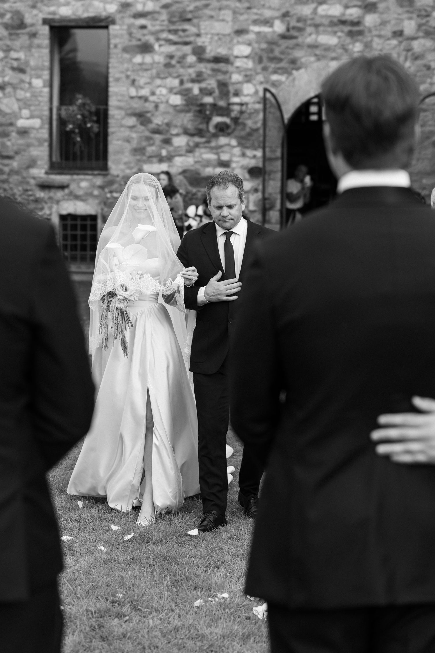 Black and white photo of a bride in veil and gown being walked to the altar by her father at an outdoor wedding.