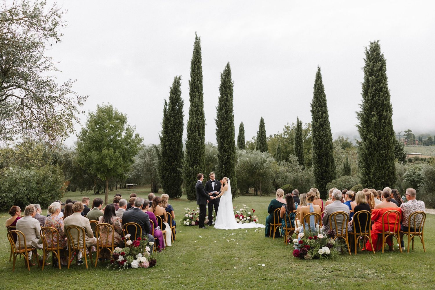Outdoor wedding ceremony on green lawn with cypress trees, guests seated in wooden chairs facing couple at altar.
