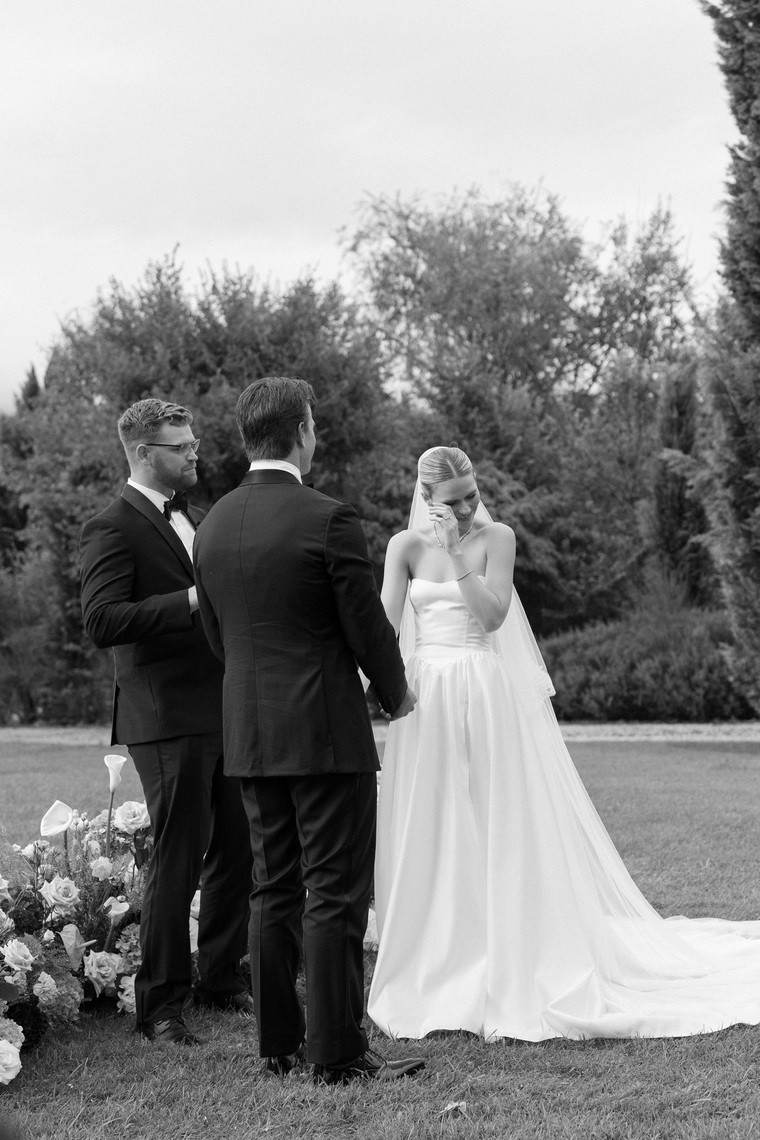 Black and white photo of a bride wiping tears during an outdoor wedding ceremony while holding hands with the groom.