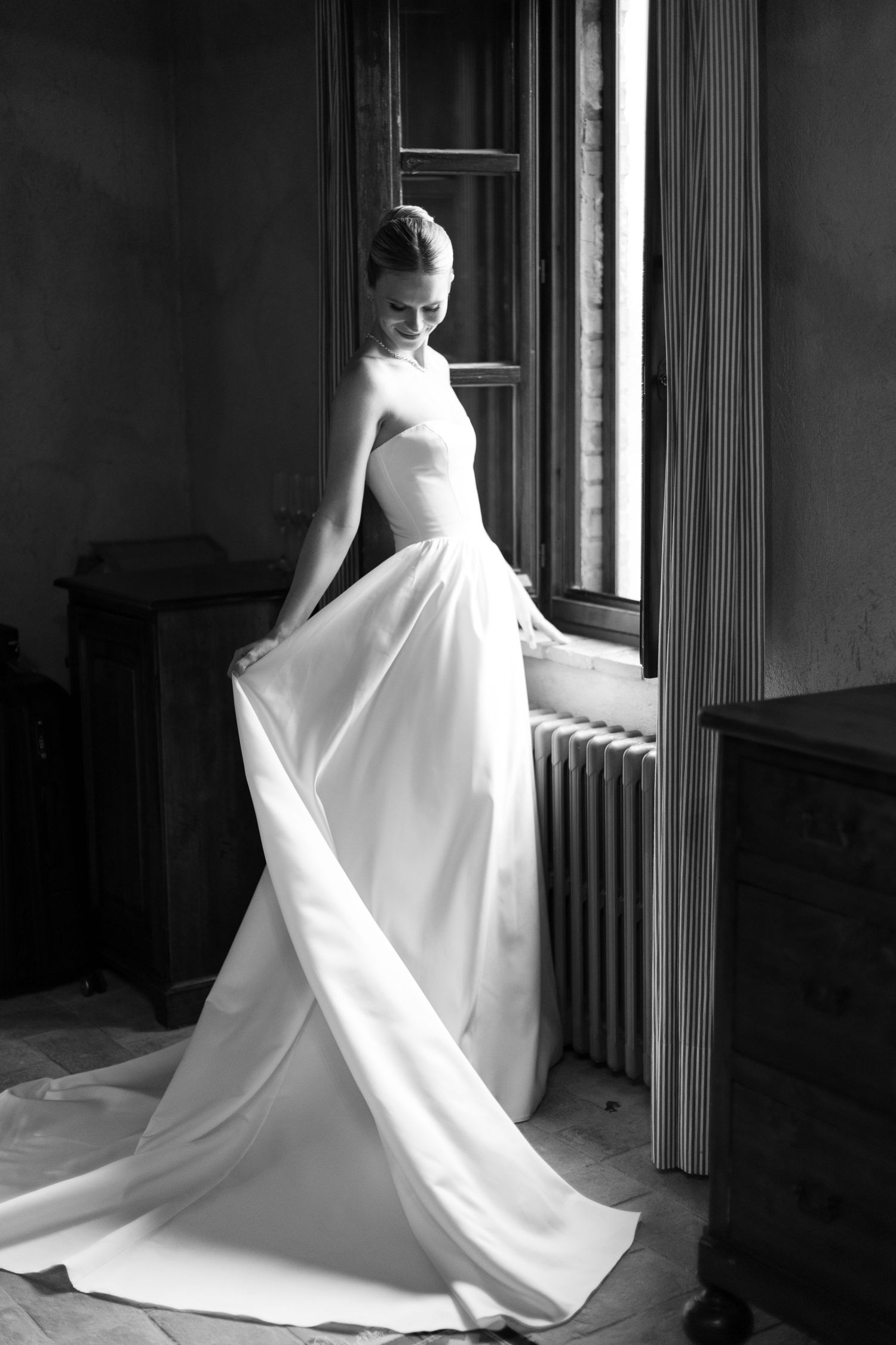 Black and white photo of a bride in a strapless flowing white gown standing by a window in an elegant room.