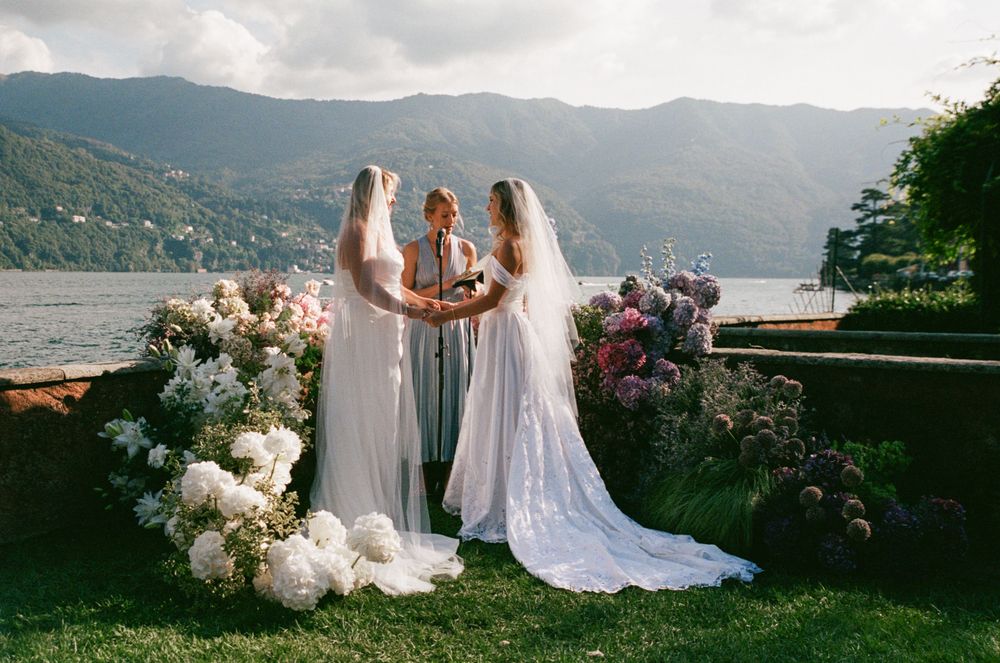 Two brides in white gowns exchanging vows at lakeside ceremony with lush floral arrangements.
