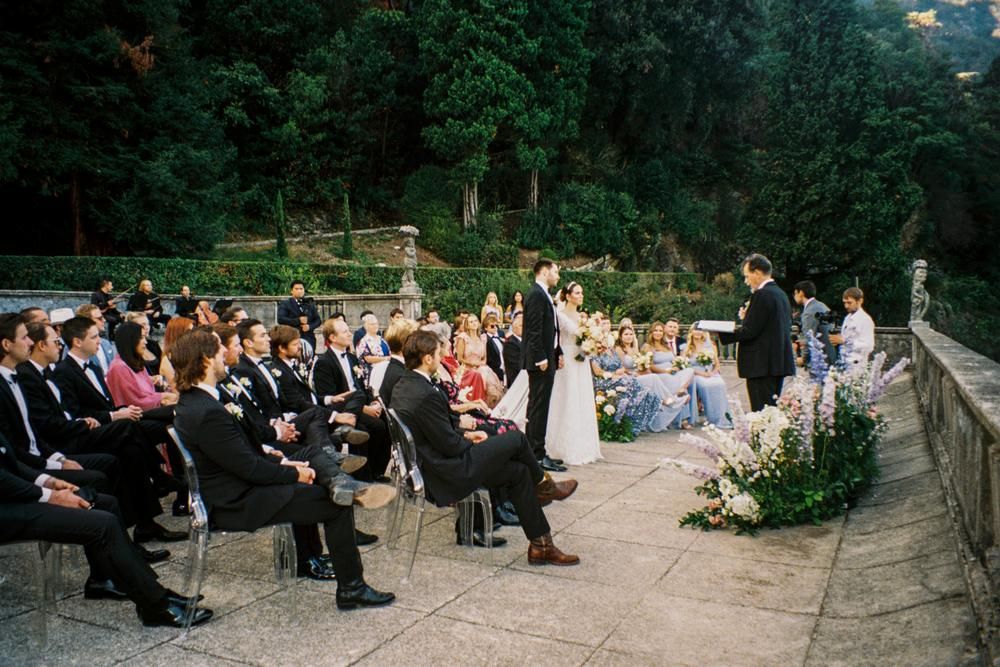 Outdoor wedding ceremony on a stone terrace with seated guests watching the couple exchange vows at dusk.