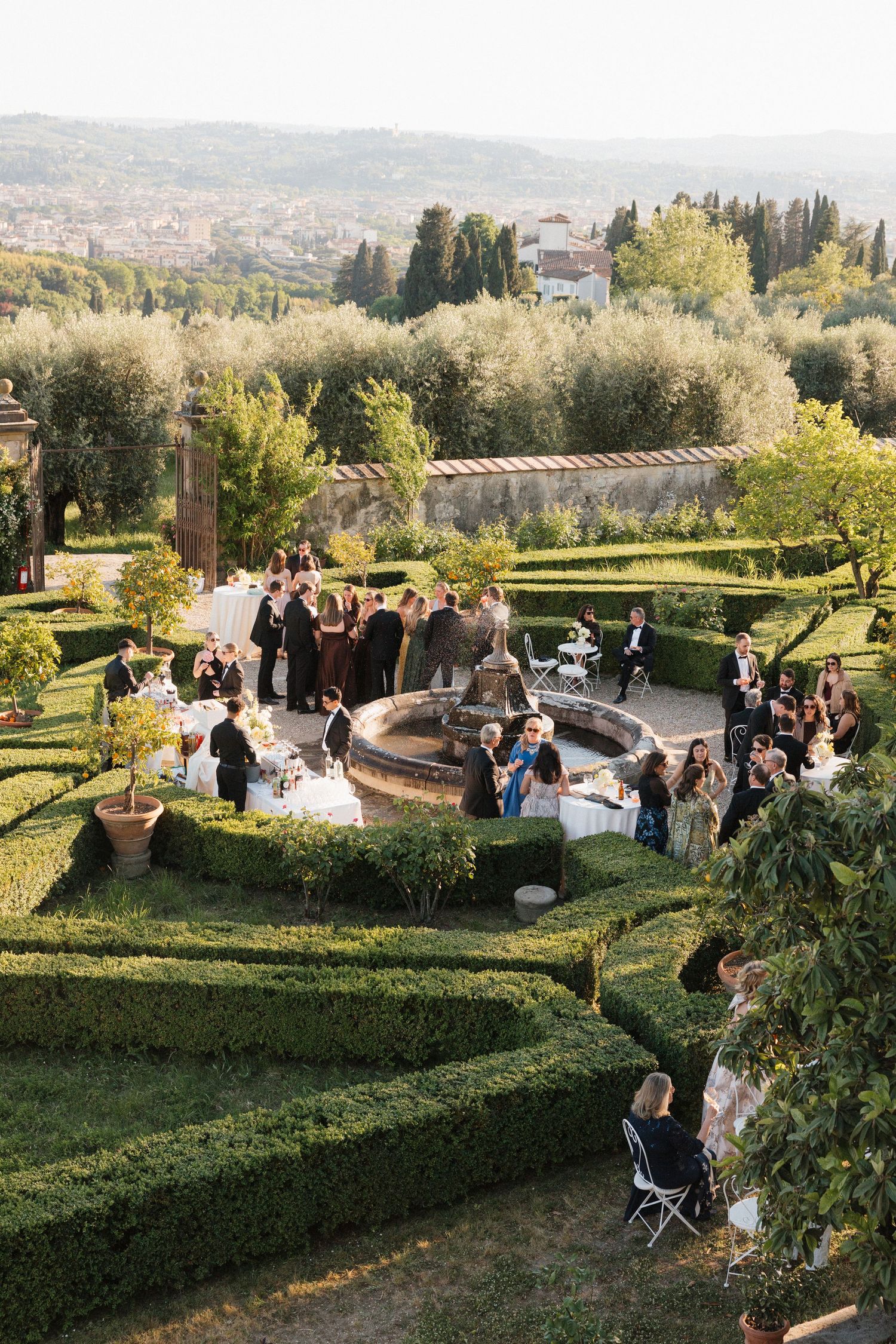 Aerial view of wedding guests mingling in an elegant Italian garden with manicured hedges and a central stone fountain.