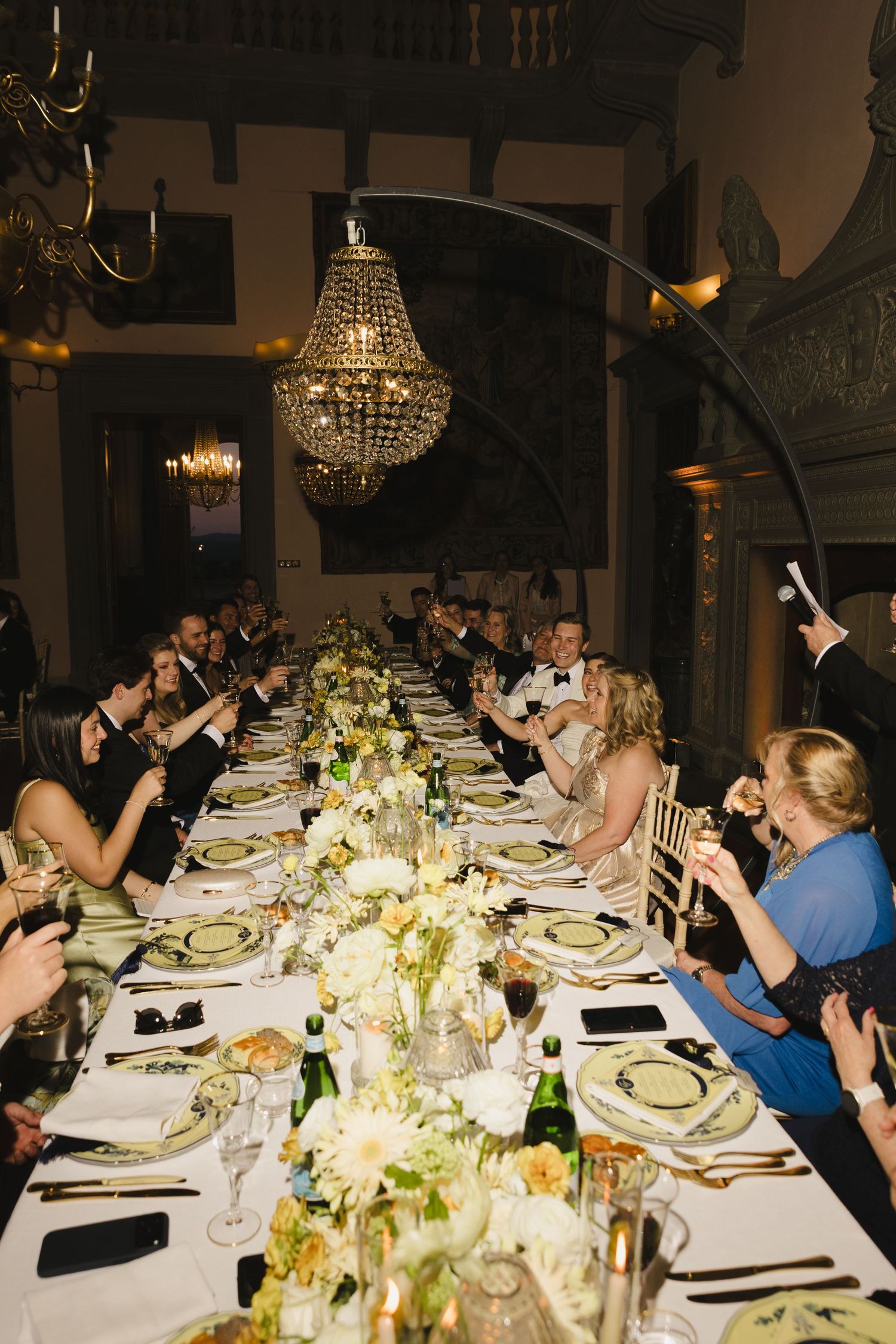 Elegant wedding reception dinner with guests raising glasses for a toast beneath a crystal chandelier in a grand hall.