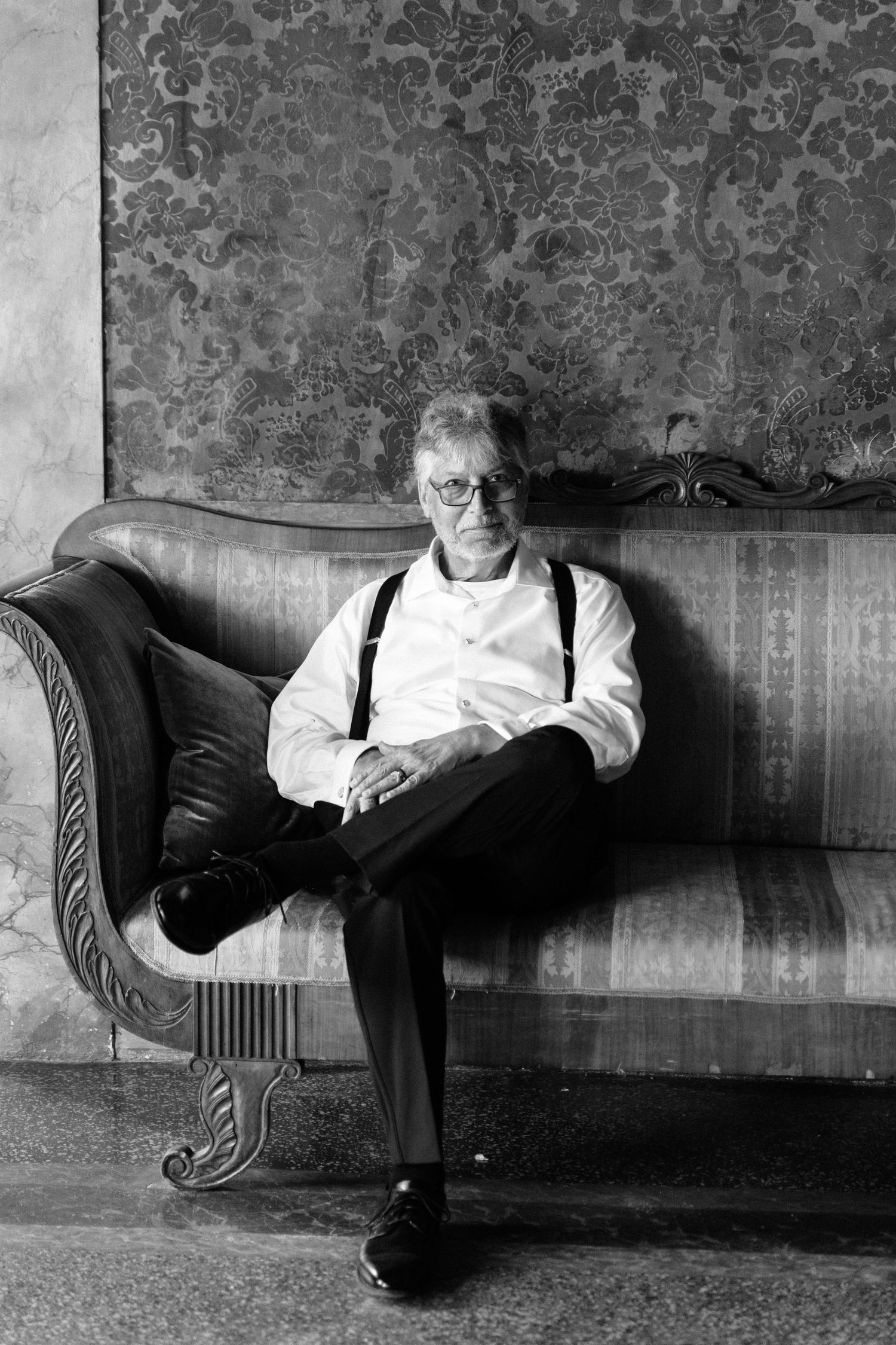 Black and white portrait of an older man in suspenders and glasses relaxing on an ornate vintage sofa.