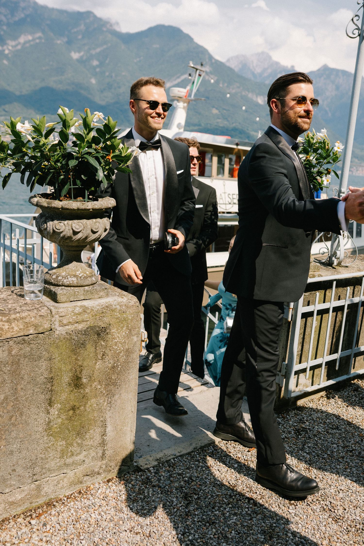 Two groomsmen in black tuxedos and sunglasses stand on a lakeside terrace with mountain views.
