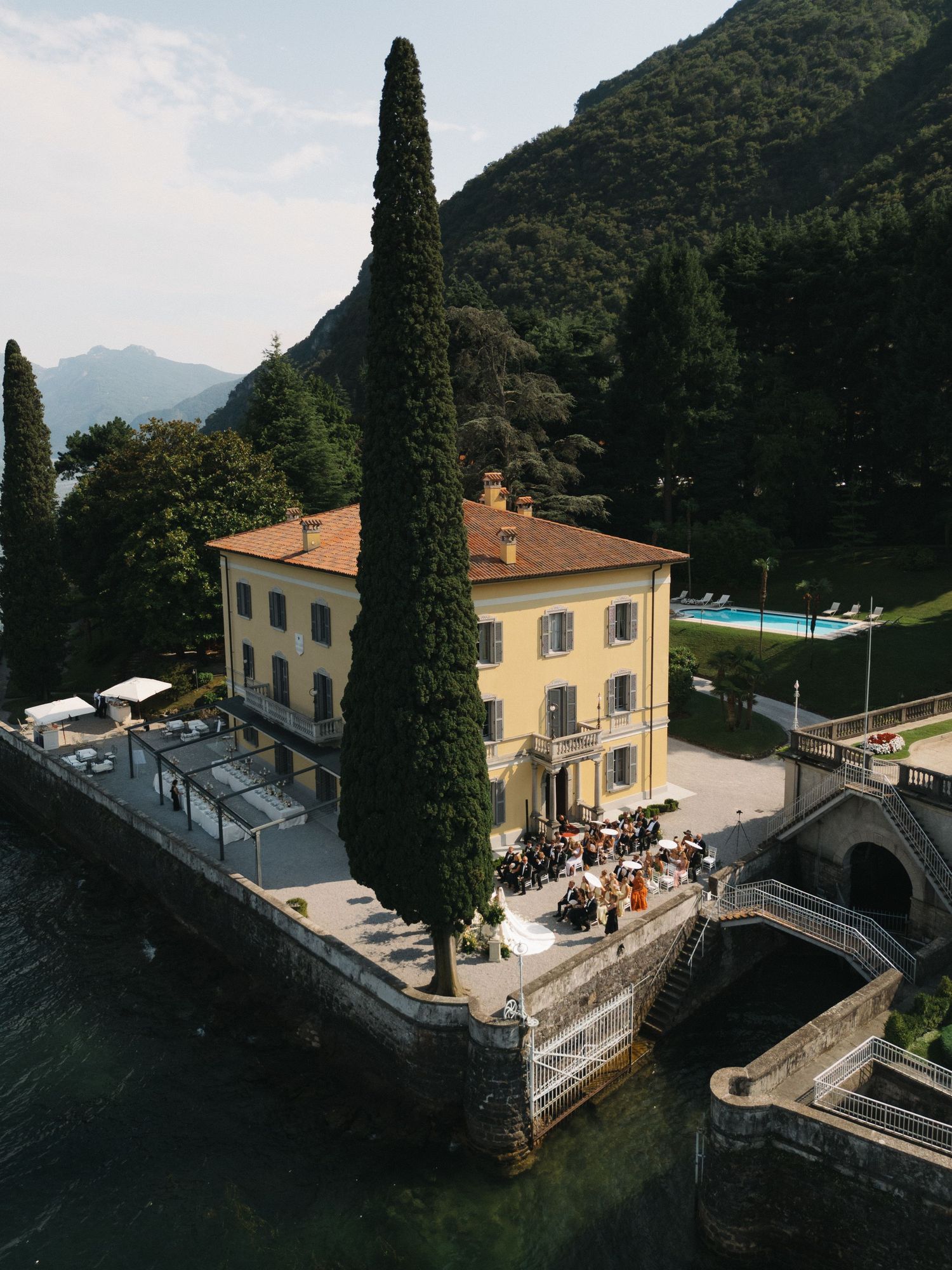 Aerial view of an elegant yellow Italian villa on Lake Como surrounded by cypress trees and mountains during a wedding celebration.