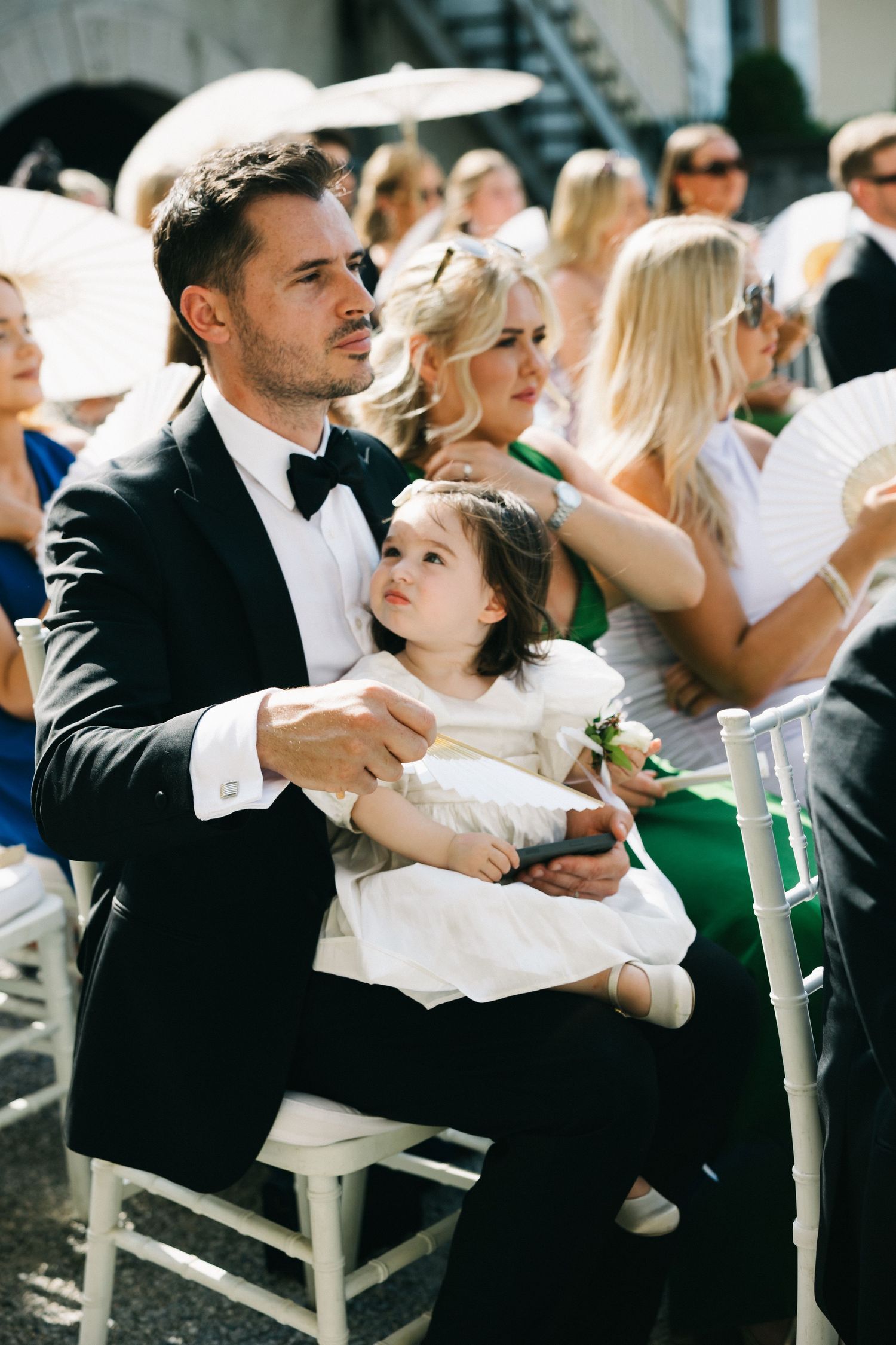 Man in tuxedo holding young girl in white dress at outdoor wedding ceremony with guests seated behind.