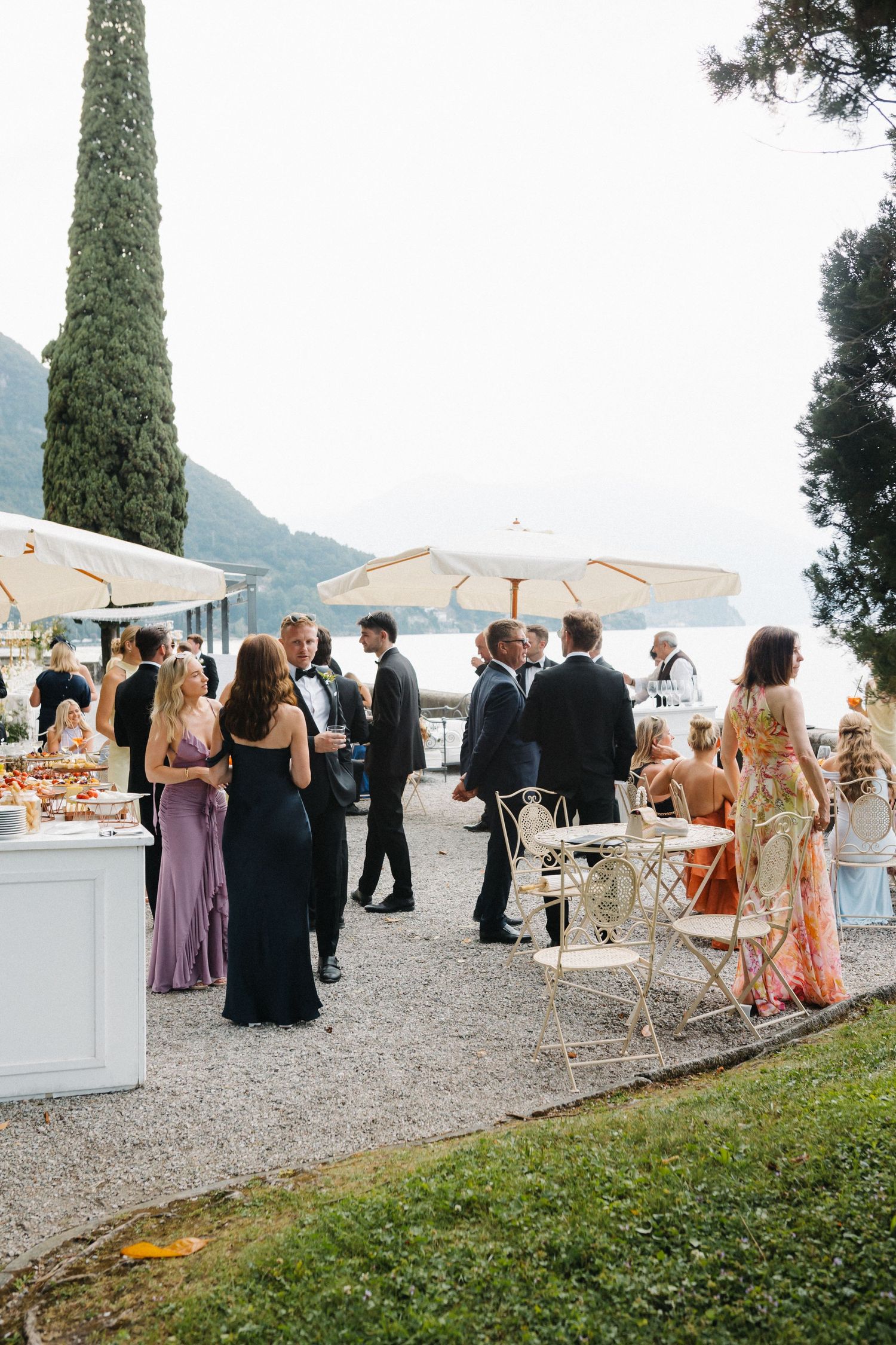 Elegant outdoor wedding cocktail hour on Lake Como terrace with guests in formal attire mingling under white umbrellas.