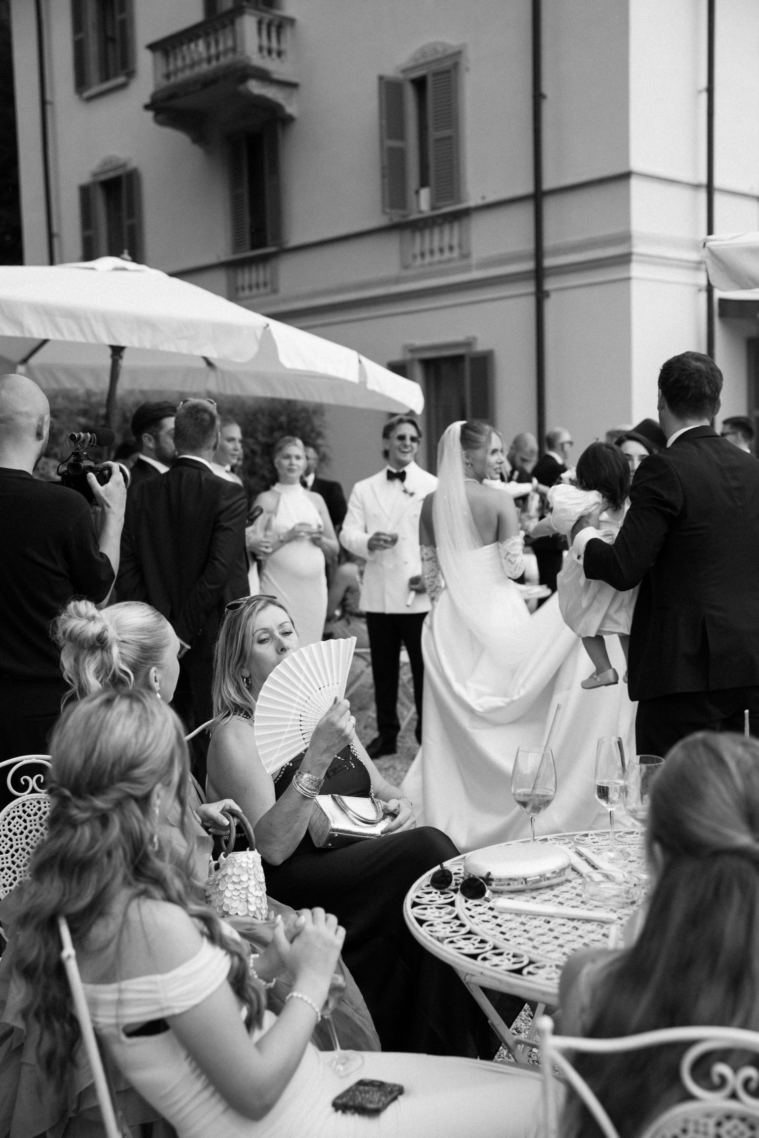 Black and white wedding reception photo with bride in veil, guests seated at ornate tables, and elegant outdoor venue.