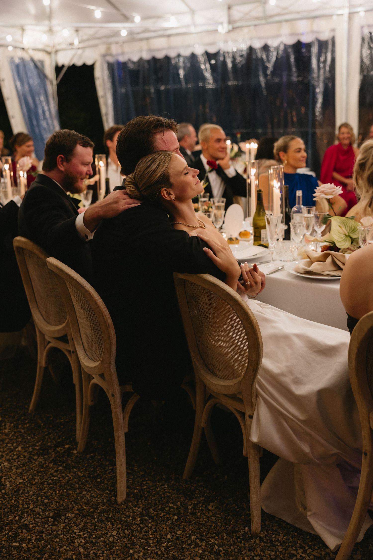 Couple embracing at elegant candlelit wedding reception dinner under a tent with guests seated nearby.