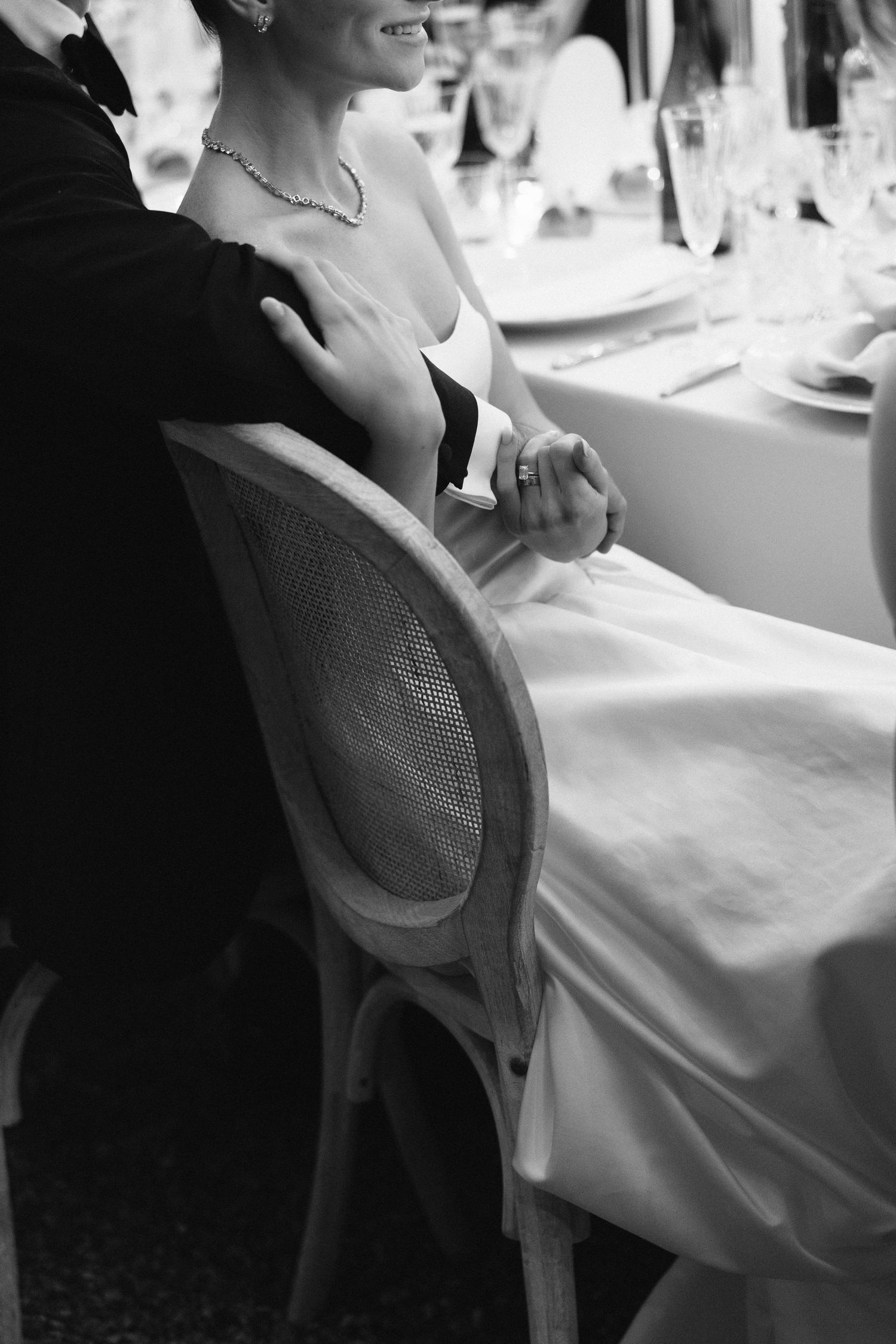 Black and white photo of bride and groom holding hands at wedding reception table, elegant formal attire.