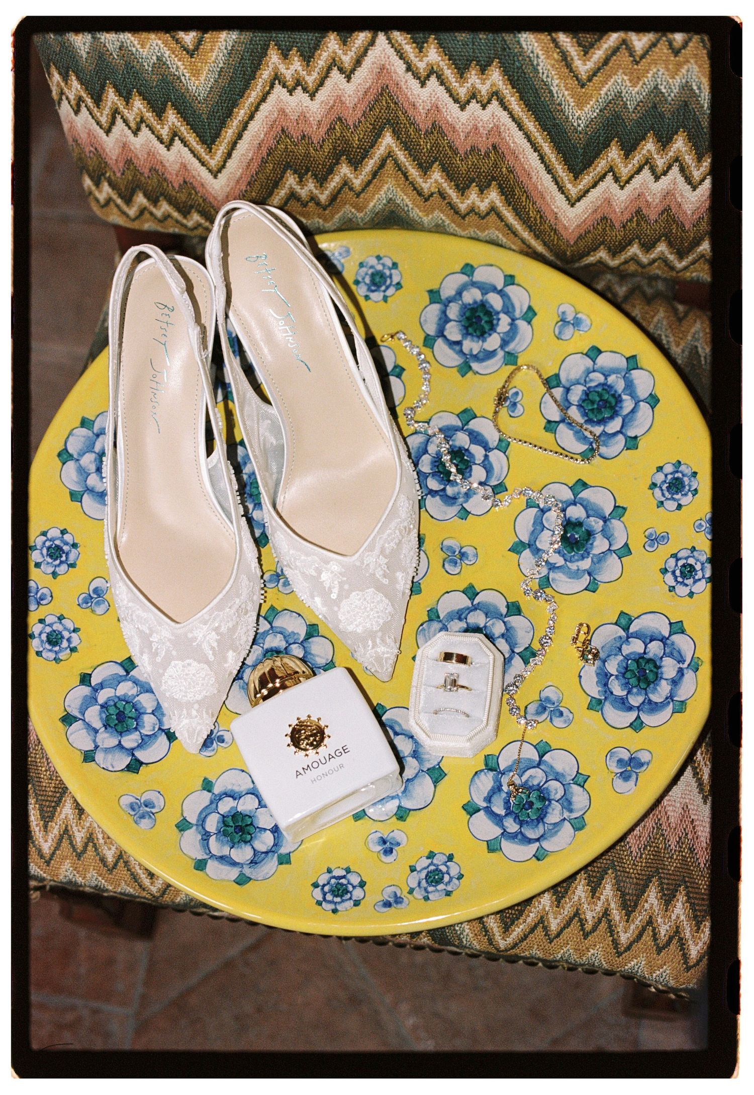 White lace bridal heels displayed on a yellow floral tray with Amouage perfume and ring box on a colorful chair.
