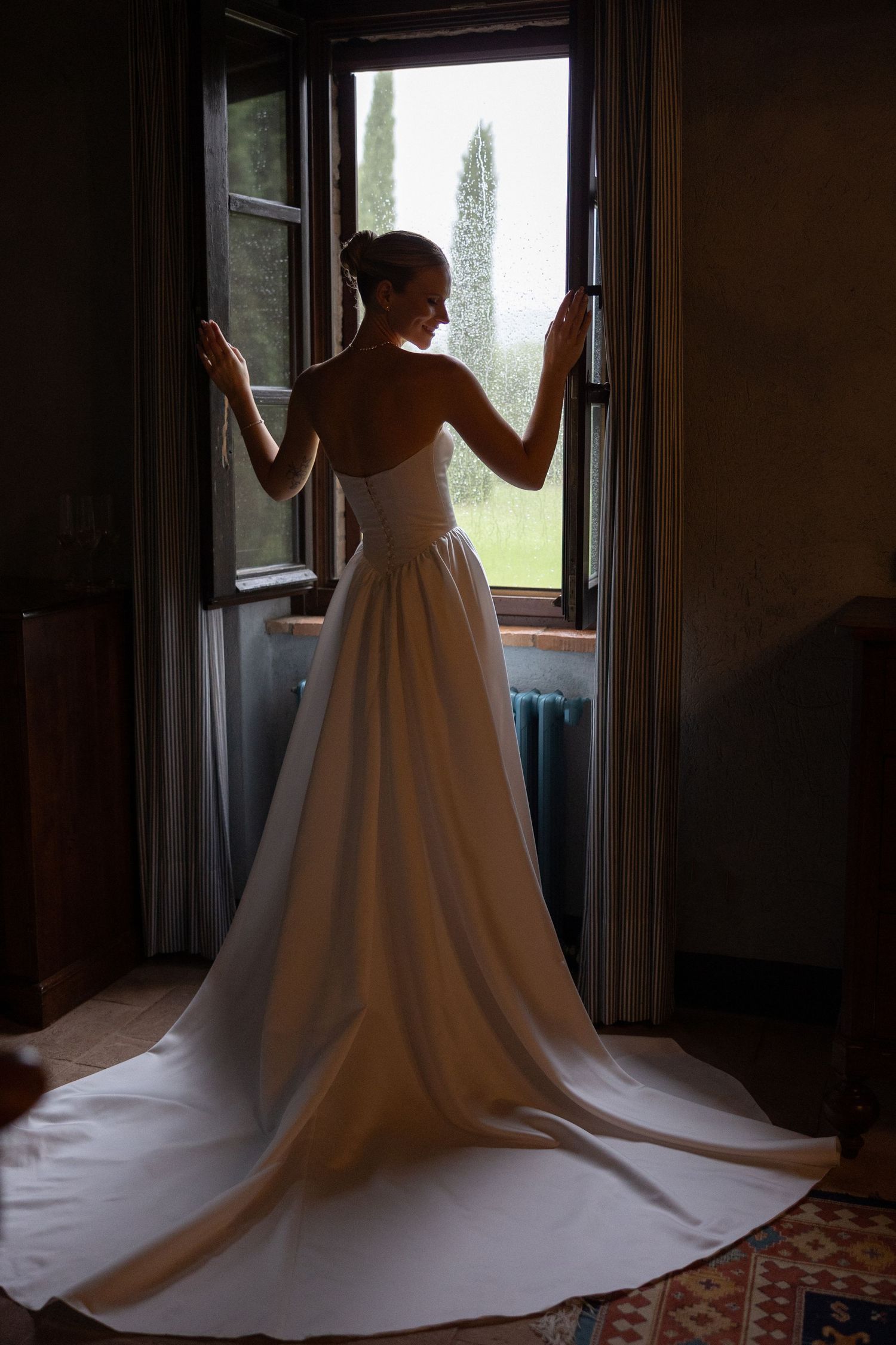 Bride in a strapless white wedding gown with a long train stands at an open window in a dimly lit room.