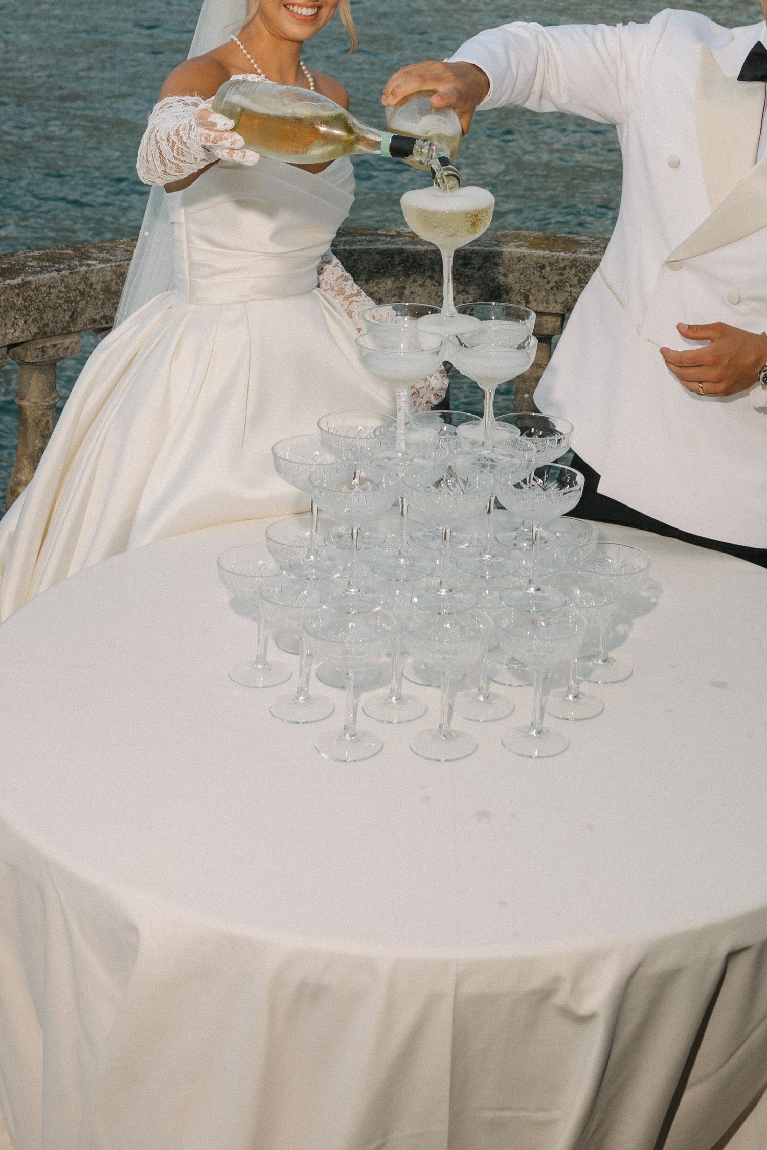 Bride and groom pouring champagne into a pyramid of glasses at an outdoor wedding reception by the water.
