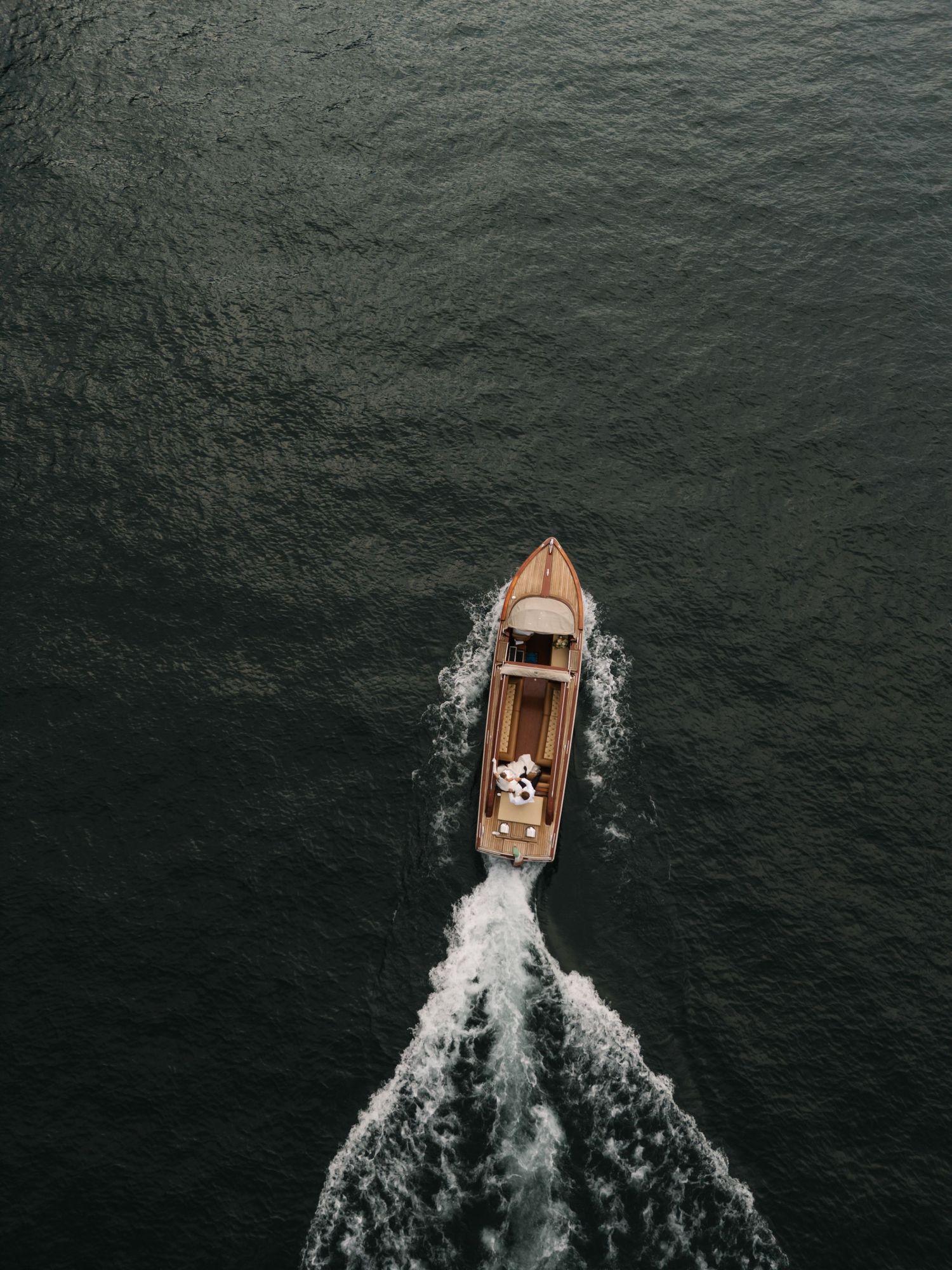Aerial view of a classic wooden speedboat cutting through dark green water, leaving a white wake trail behind.