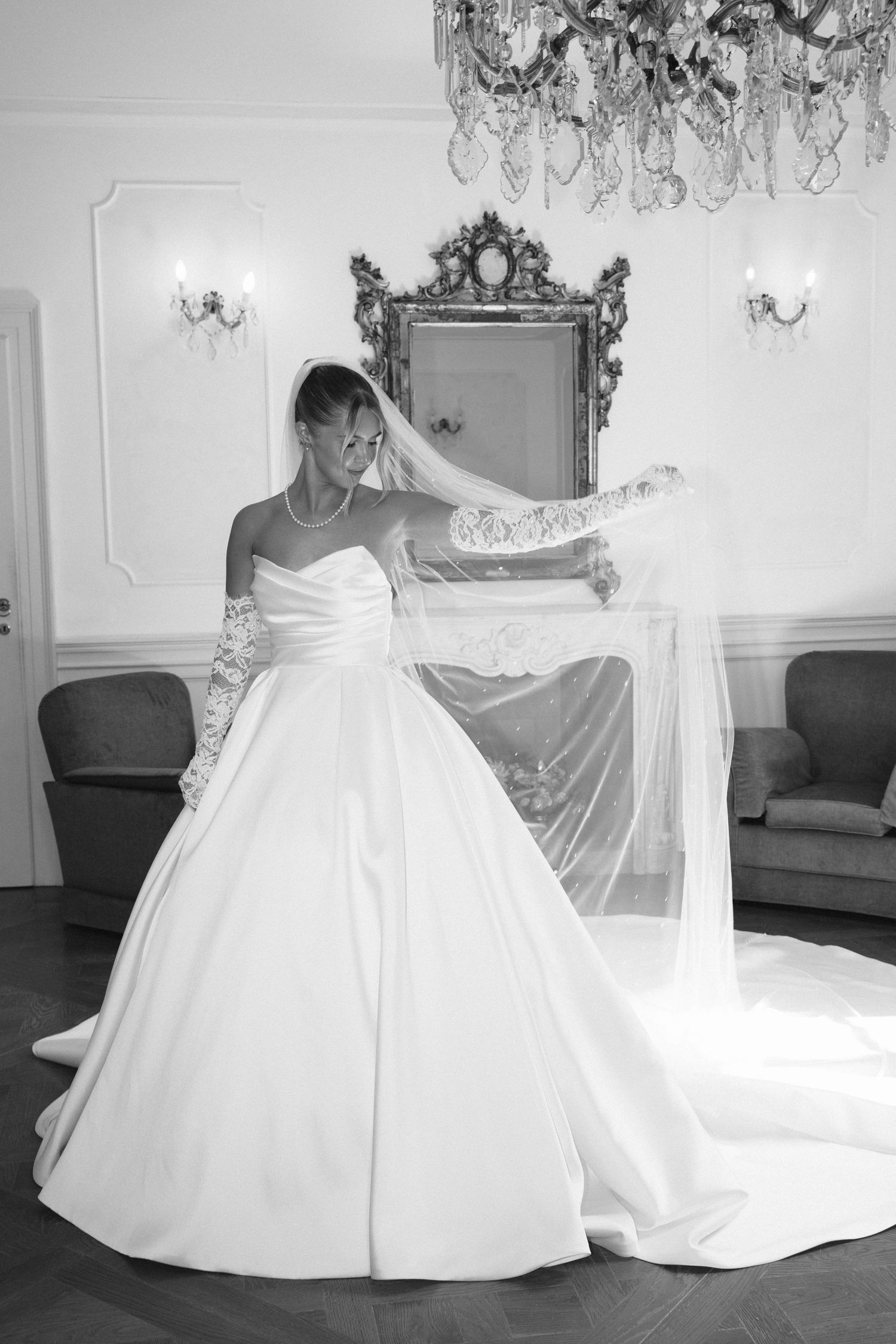 Elegant bride in a white ball gown with veil and lace gloves poses in a grand room with crystal chandelier.