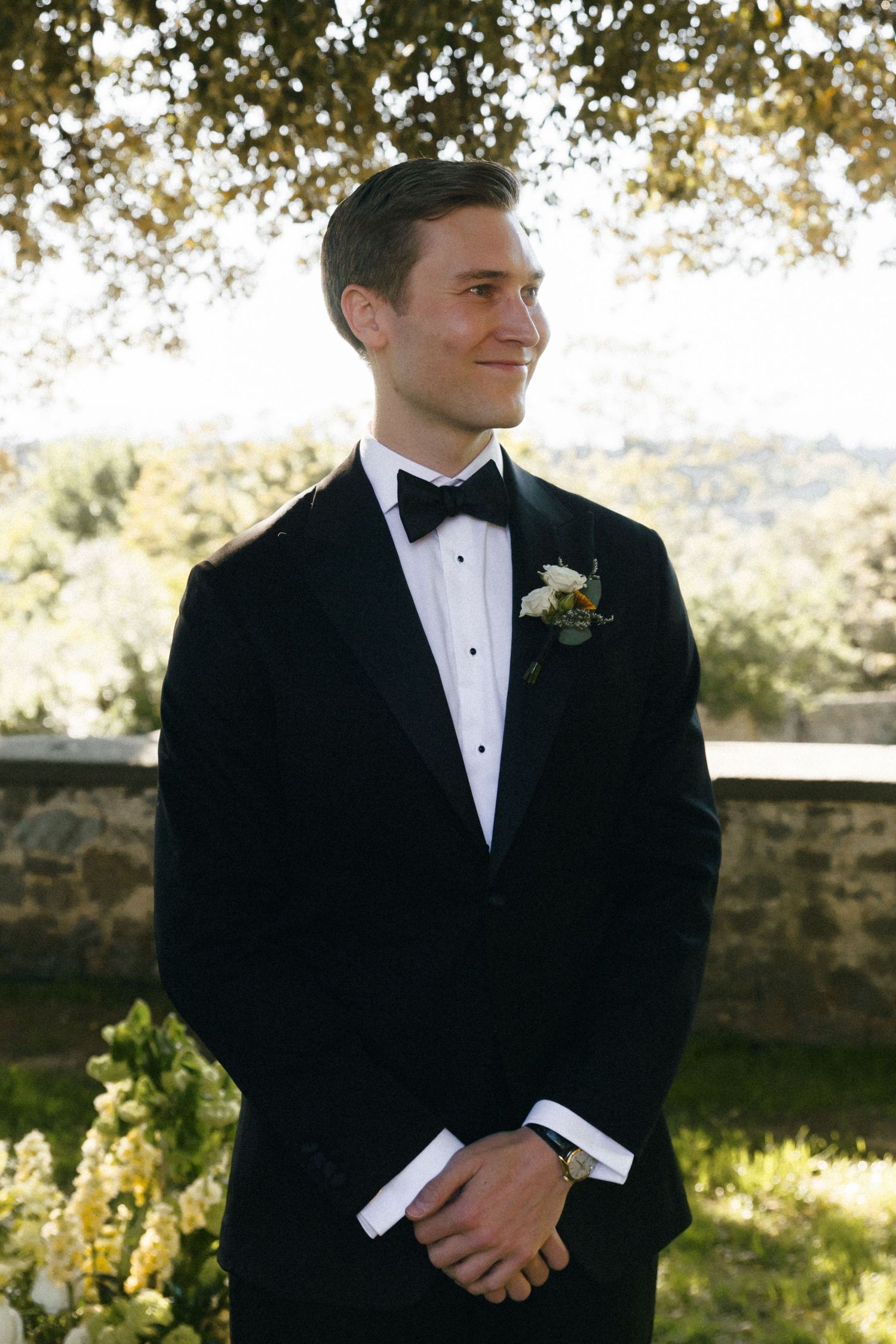 Elegant groom in black tuxedo with boutonniere smiling outdoors at wedding ceremony.