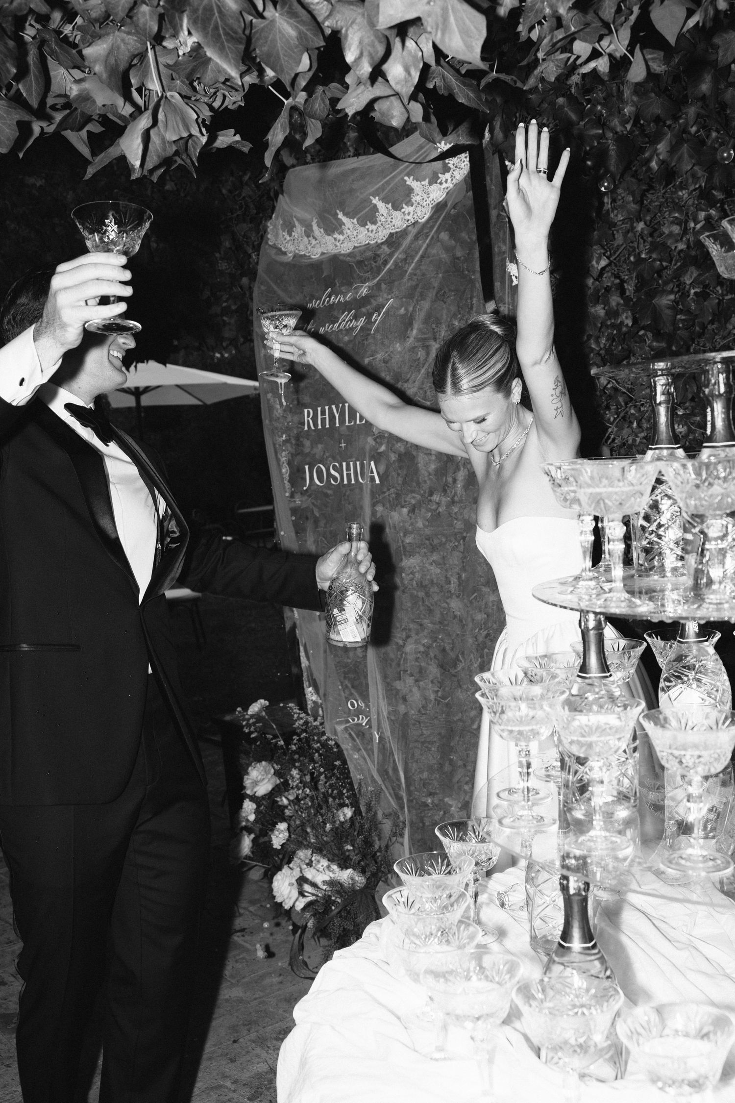 Black and white photo of bride and groom celebrating at champagne tower during wedding reception.