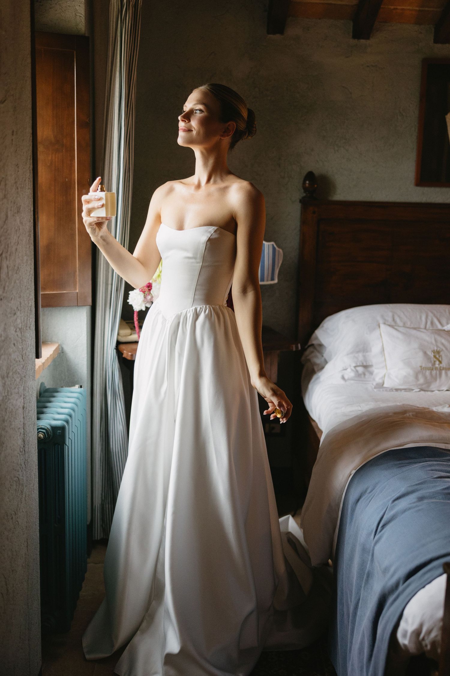Bride in strapless white wedding gown holding champagne glass by window in rustic bedroom.