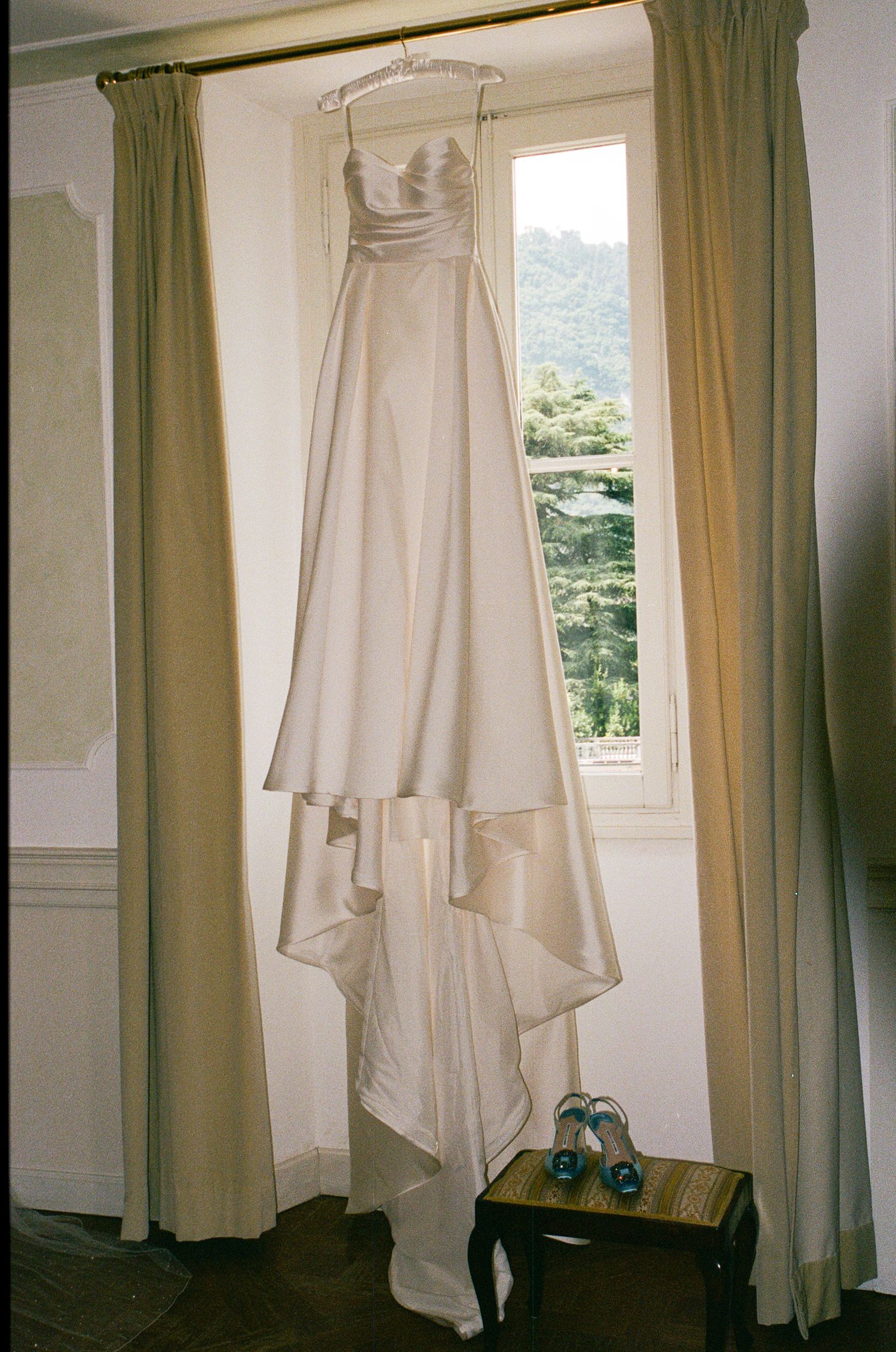 Elegant strapless ivory wedding gown hanging by a window with blue bridal shoes on a wooden stool below.