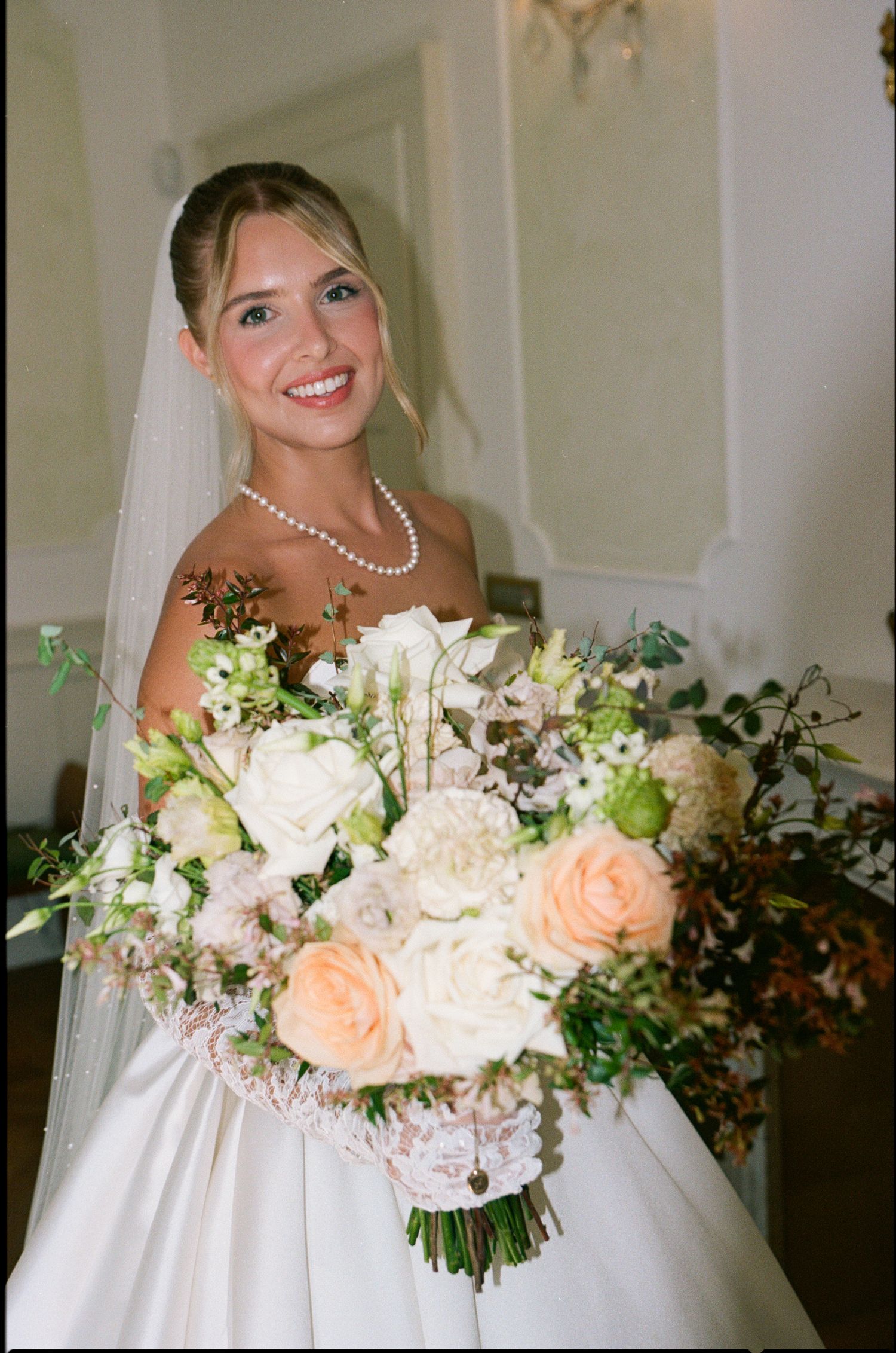 Smiling bride in white gown and veil holding lush bouquet of peach and white roses with greenery, wearing pearl necklace.