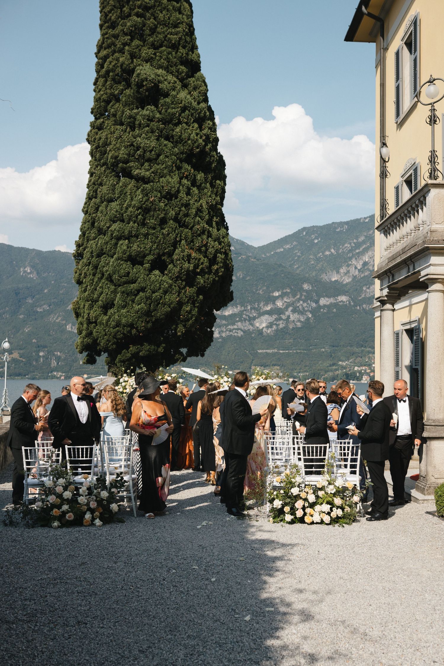 Elegant outdoor wedding ceremony at Lake Como villa with guests in formal attire, floral arrangements, and cypress tree backdrop.
