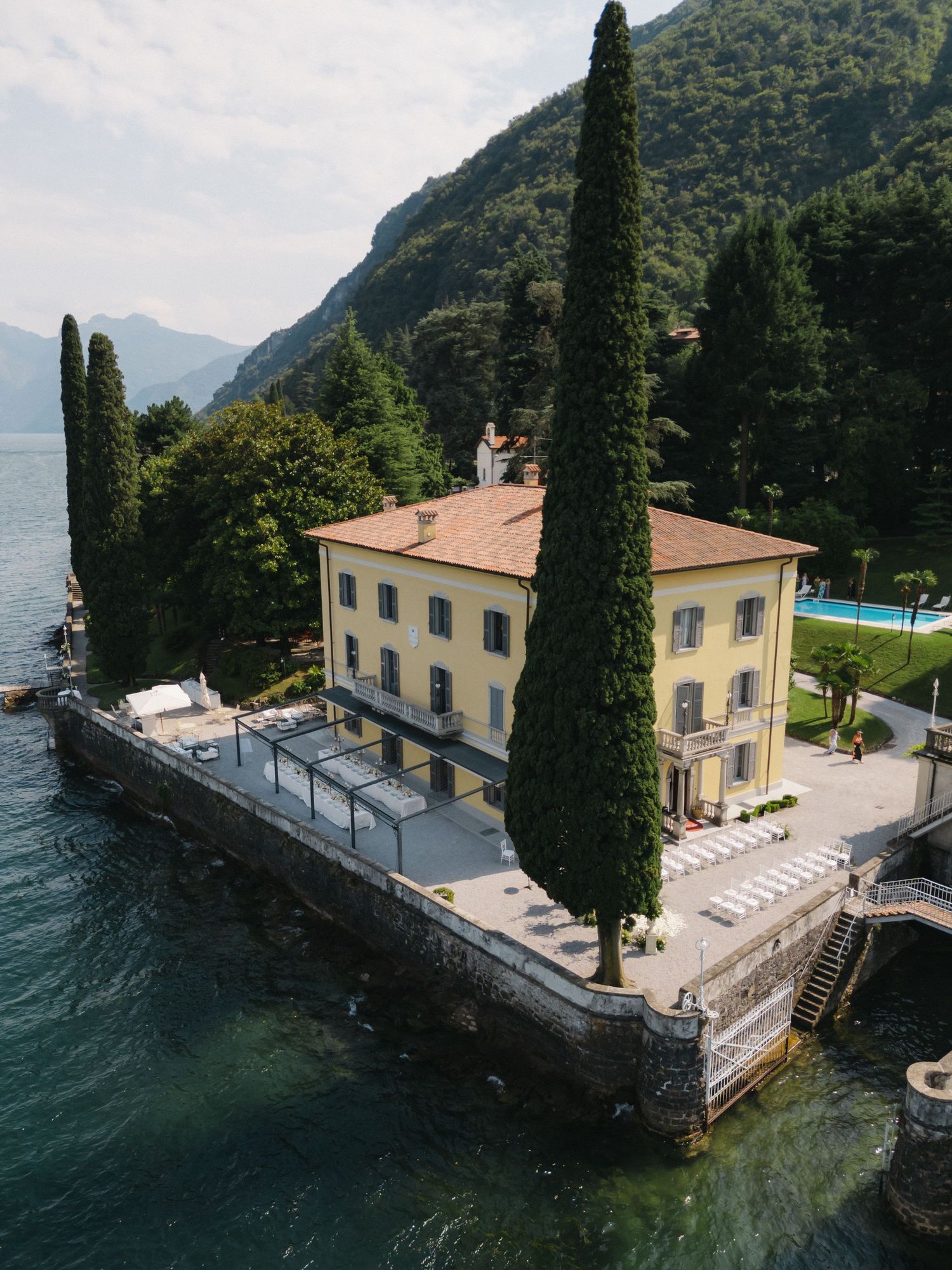 Aerial view of a yellow lakeside villa with cypress trees on Lake Como, Italy, surrounded by mountains.