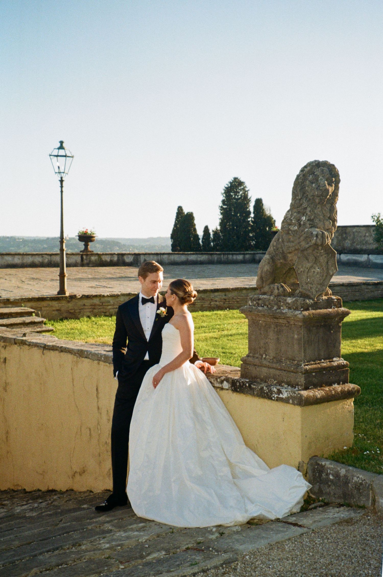 Bride and groom pose by a stone lion statue at an elegant outdoor Italian villa wedding venue.