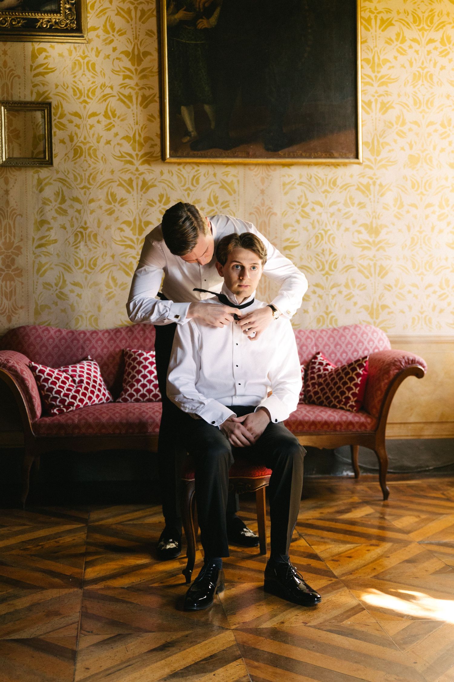 Man helping groom adjust bow tie while seated on chair in elegant room with ornate wallpaper and vintage sofa.