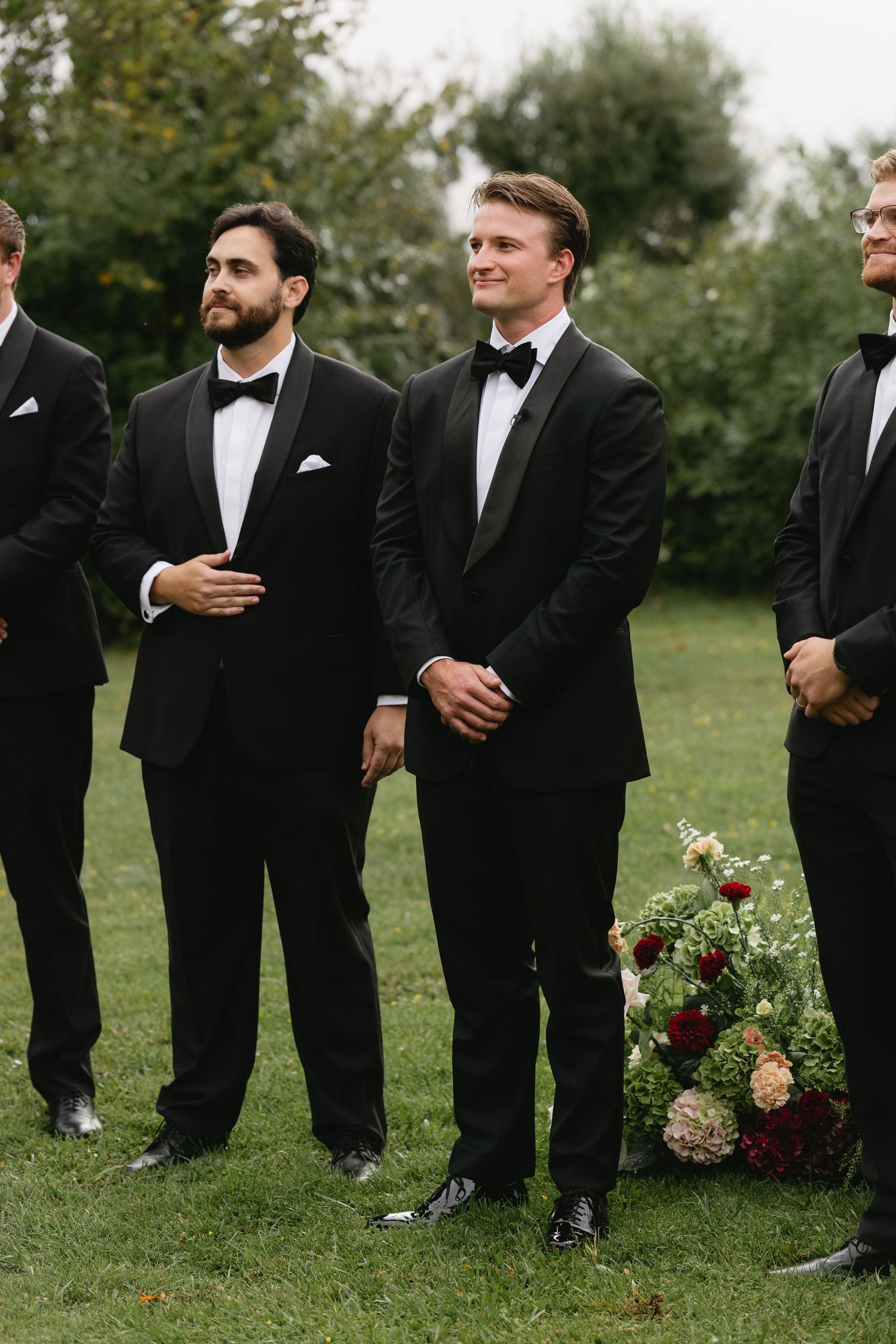 Groomsmen in black tuxedos with bow ties stand outdoors at a wedding ceremony near a floral arrangement.