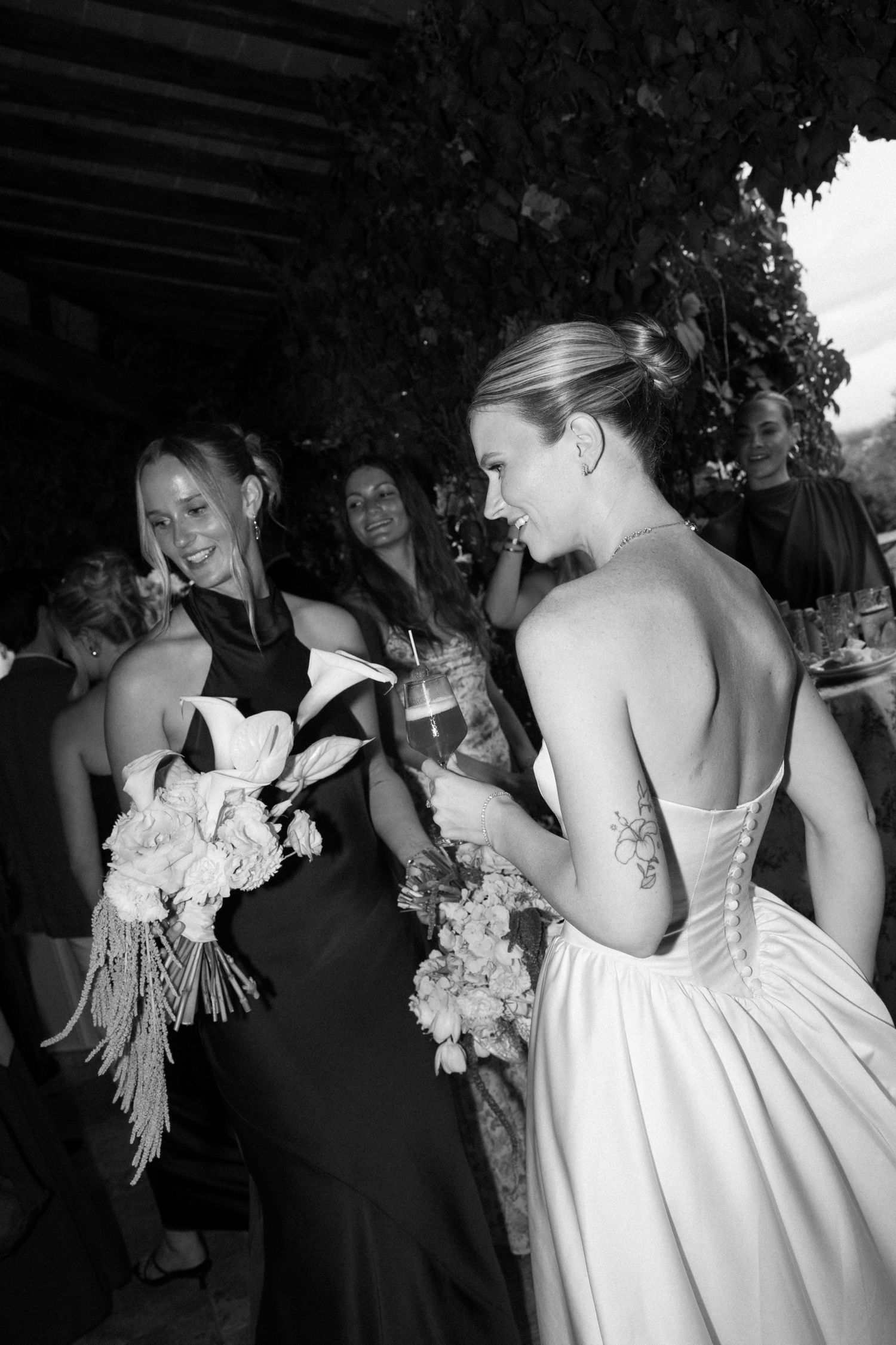 Black and white photo of bride in strapless gown with tattoo chatting with bridesmaid holding bouquet at wedding reception.
