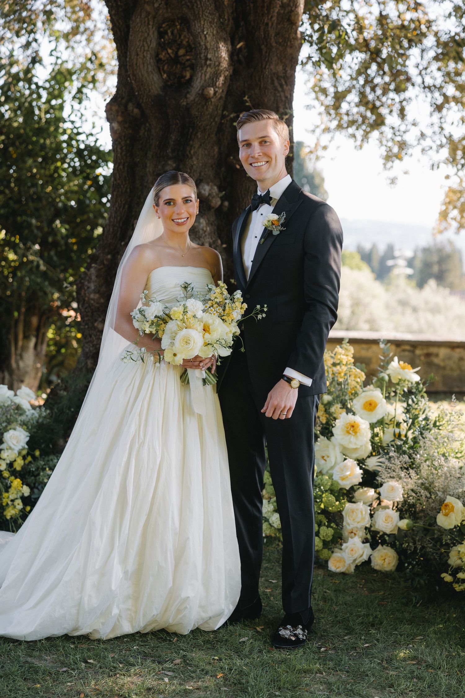 Bride and groom pose together outdoors under a large tree, surrounded by white and yellow floral arrangements.