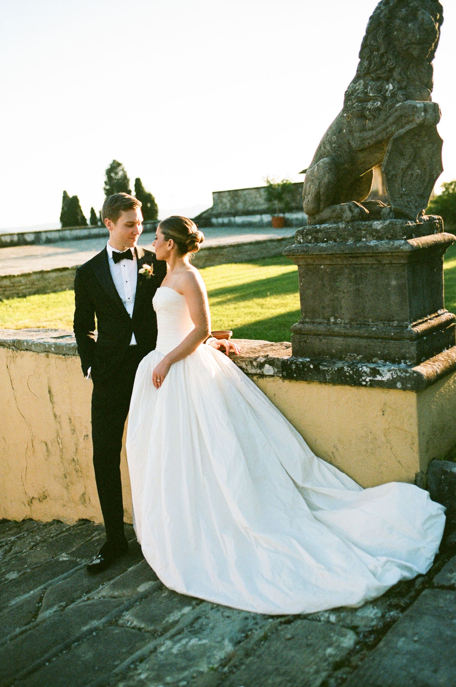 Bride in white ballgown and groom in tuxedo share romantic moment on stone terrace with lion statue.