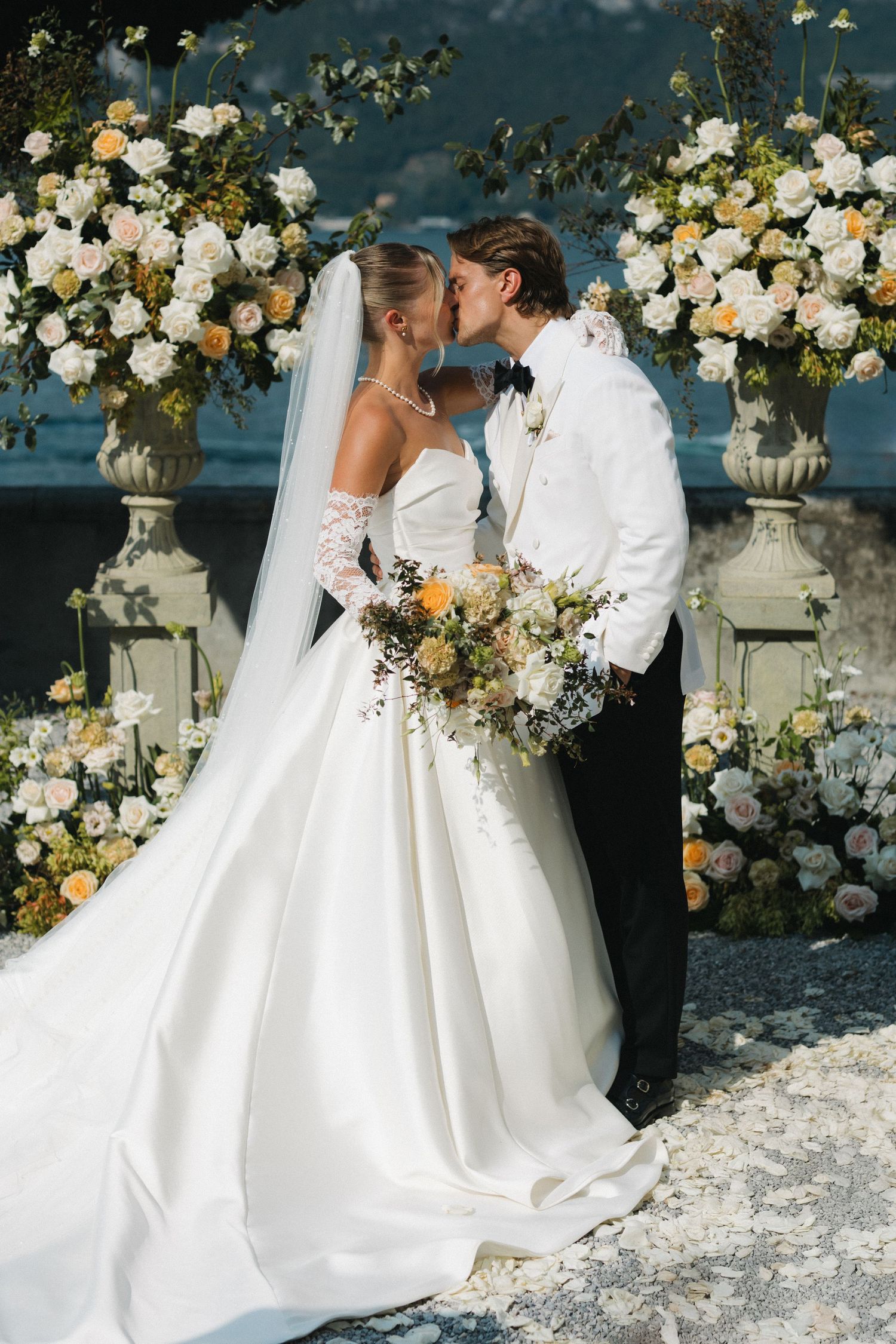 Bride and groom share a kiss at floral altar with white and peach roses, bride in strapless gown with veil and bouquet.