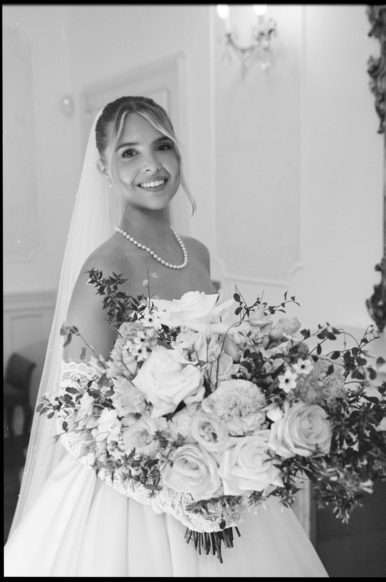 Smiling bride in white gown holds large rose bouquet, wearing pearl necklace and veil, black and white wedding photo.