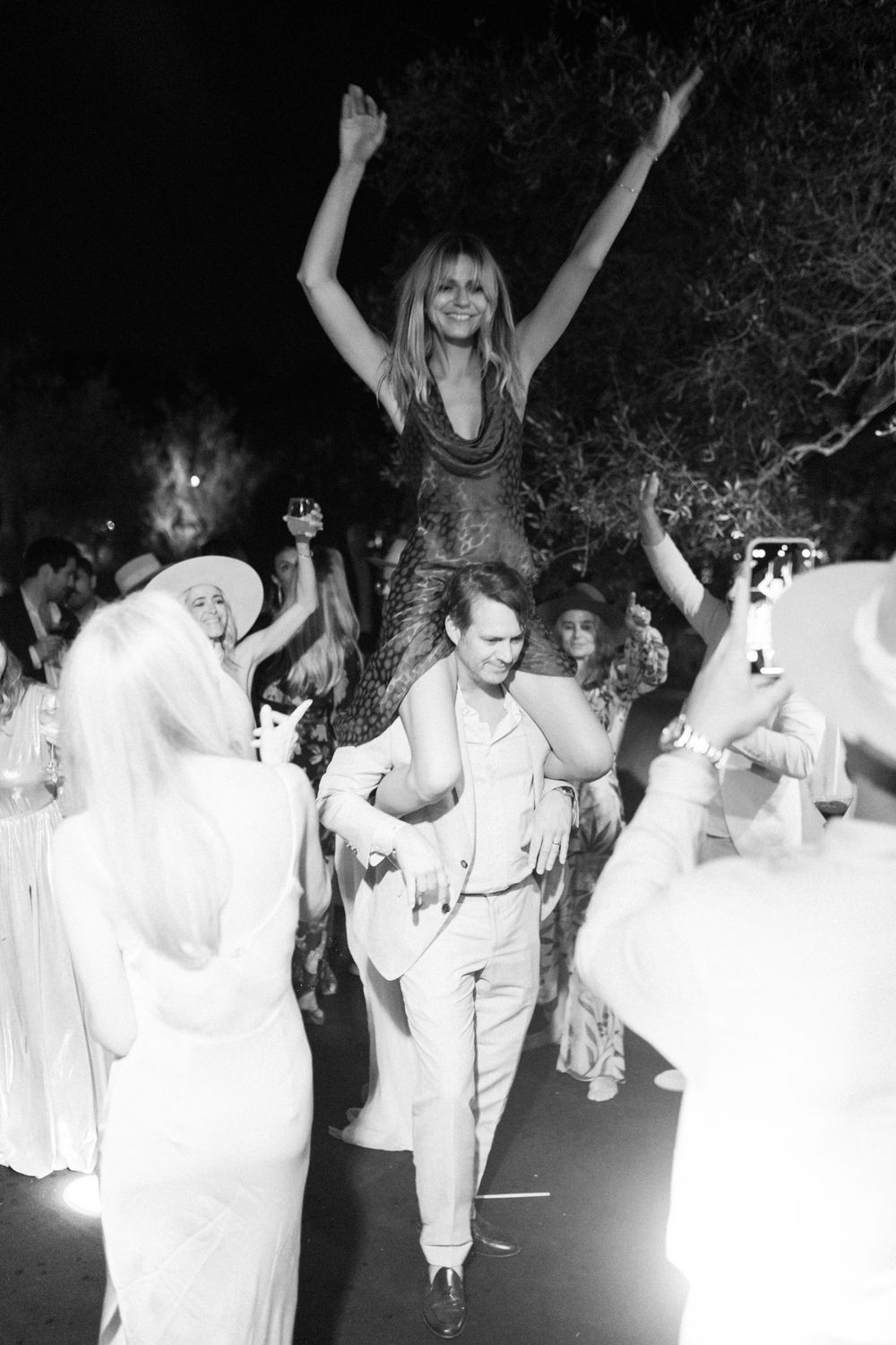 Black and white photo of a woman with arms raised being carried on shoulders at a festive outdoor night celebration.