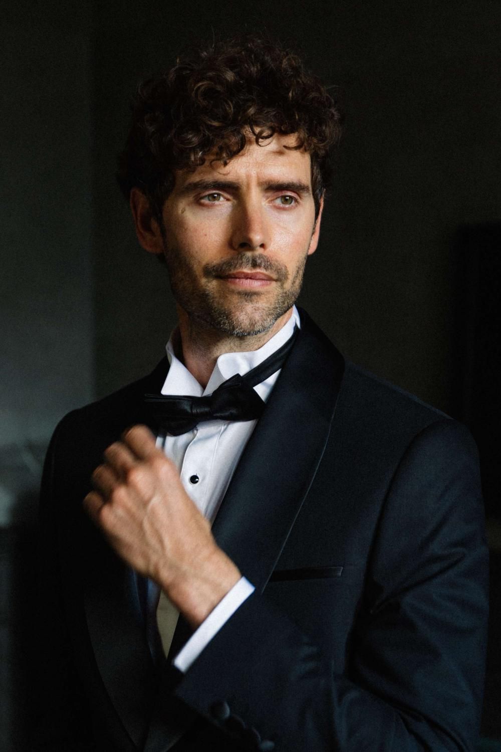 Handsome groom with curly hair adjusting his black bow tie tuxedo in dramatic moody lighting.