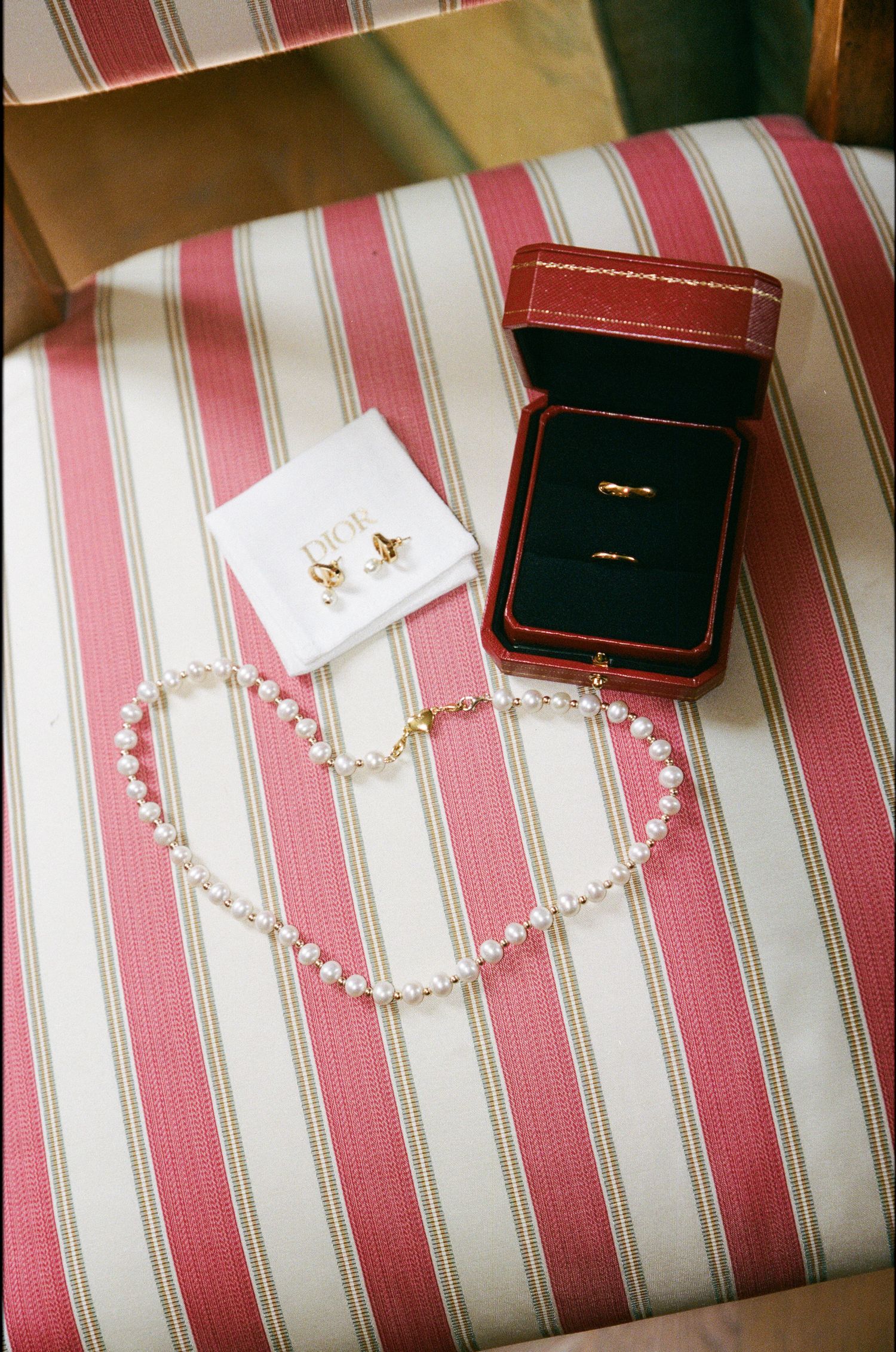 Pearl necklace, gold earrings, and two gold wedding bands in a red Cartier box displayed on a striped chair.
