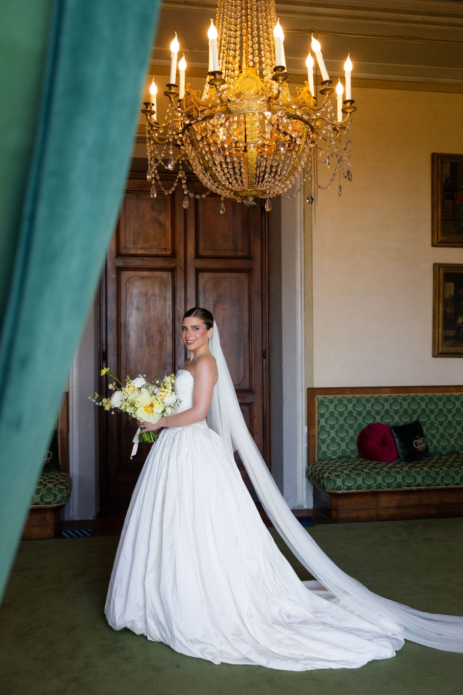 Bride in a white gown with veil holds bouquet under a golden chandelier in an elegant green room.